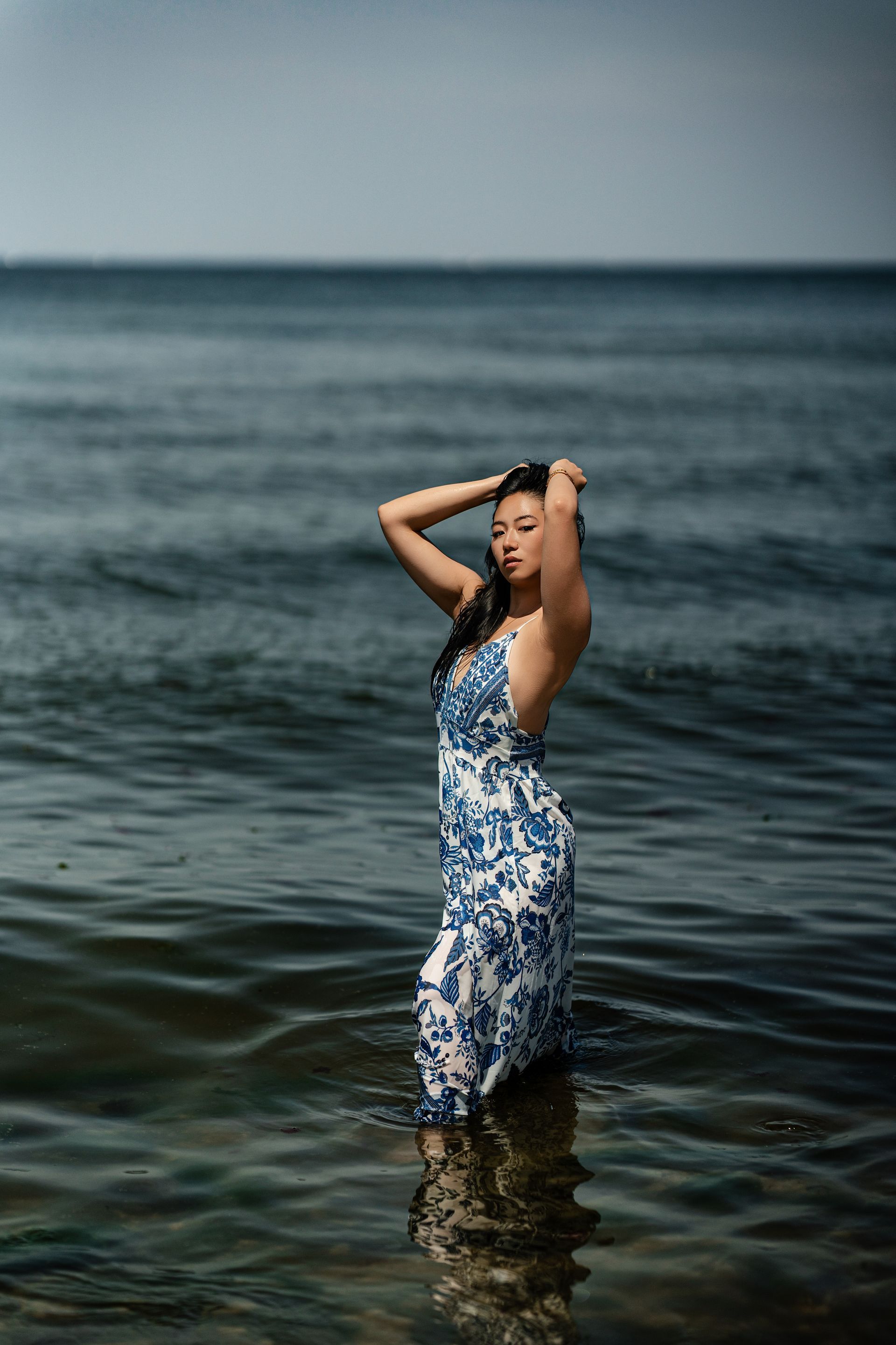 Woman in blue and white dress in the ocean with hands on head, ocean backdrop.