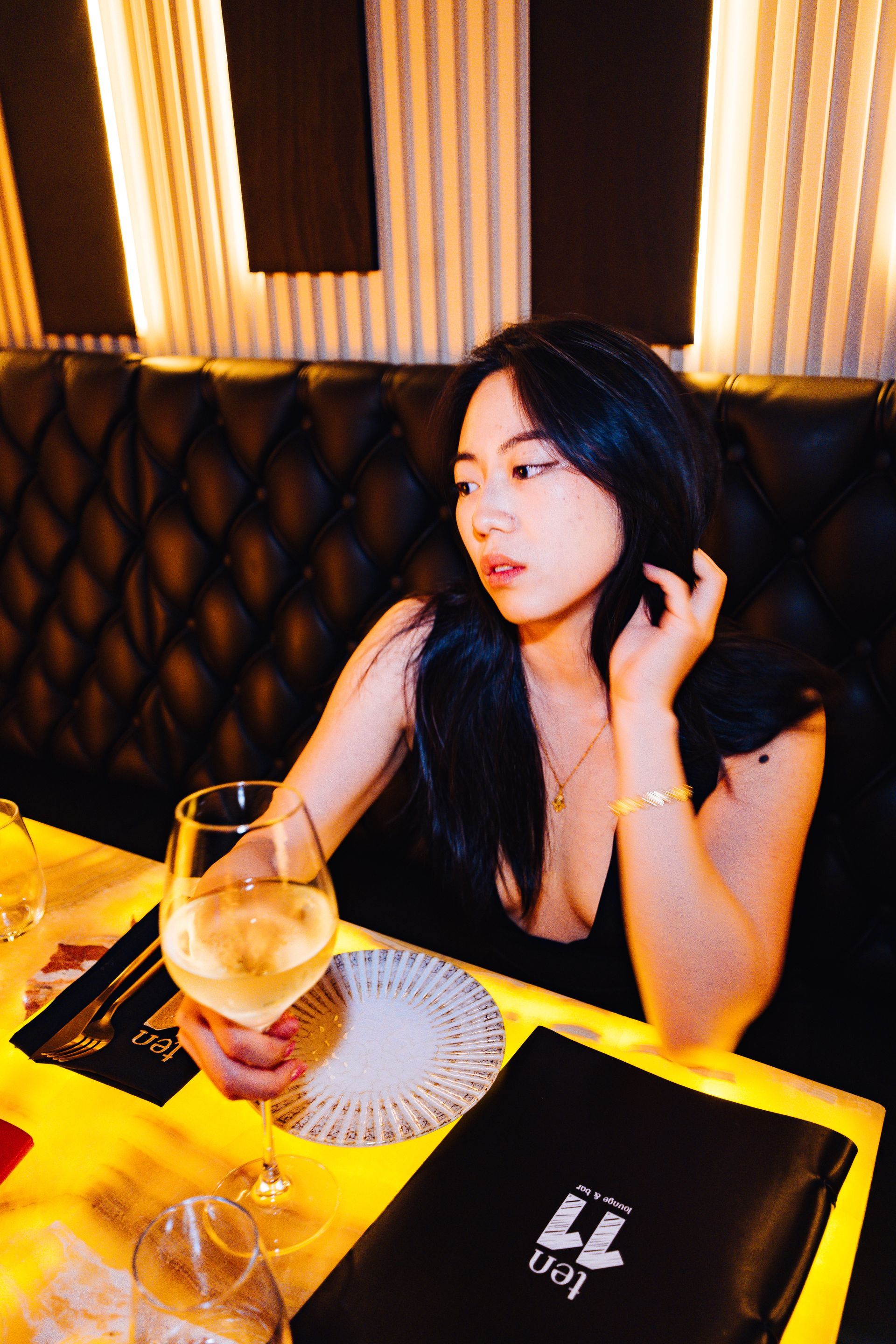 Woman seated at a table in a restaurant, holding a glass of wine. Black dress, gazing away.