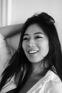 Smiling Asian woman with long hair, wearing a white shirt, leaning back indoors. Black and white headshot. 