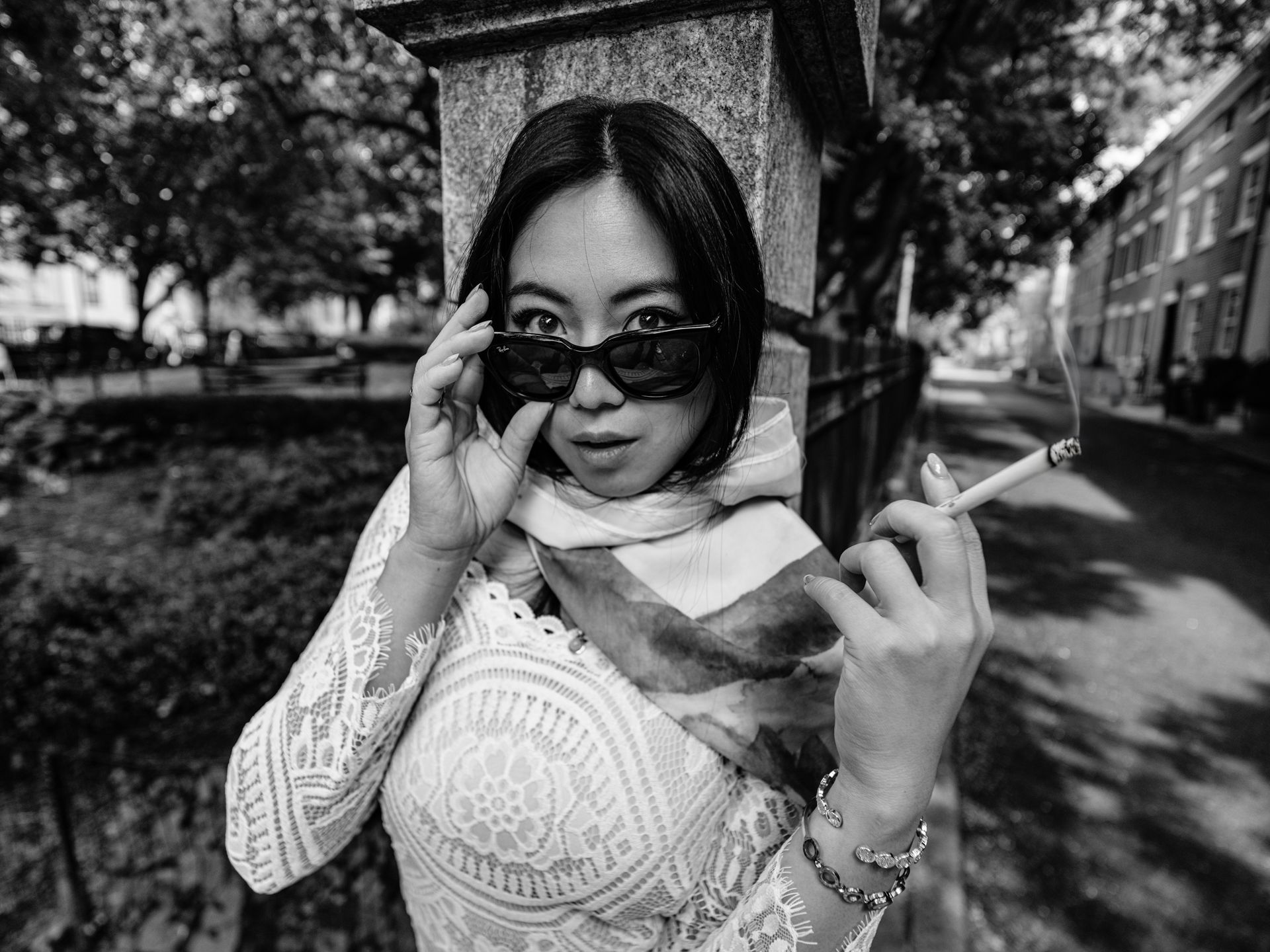 Woman in sunglasses, holding cigarette, peeking over glasses. Wearing lace top, scarf, and bracelets. Leaning against pillar outdoors.