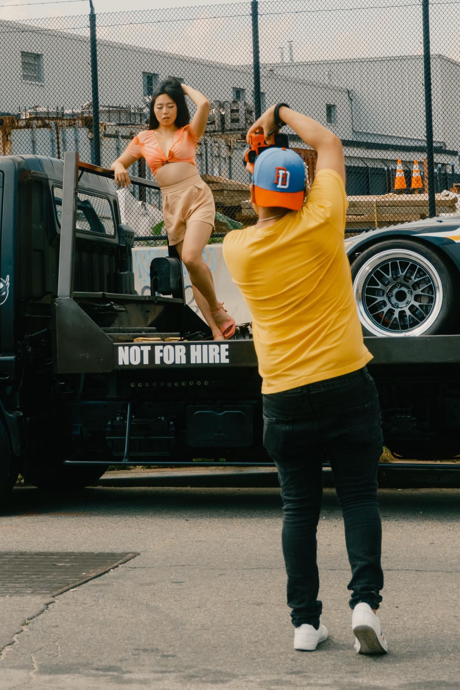 A person photographs a woman posing on a tow truck for Crown Speed Auto Block Party. Car model. The setting is outdoors.