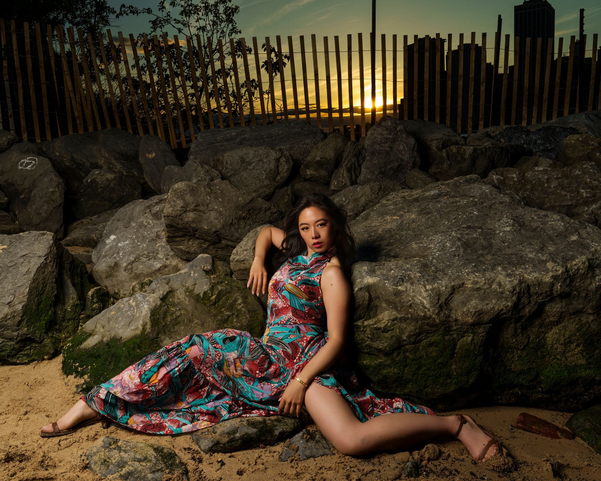 Woman in floral dress reclines on rocks by the beach at sunset.