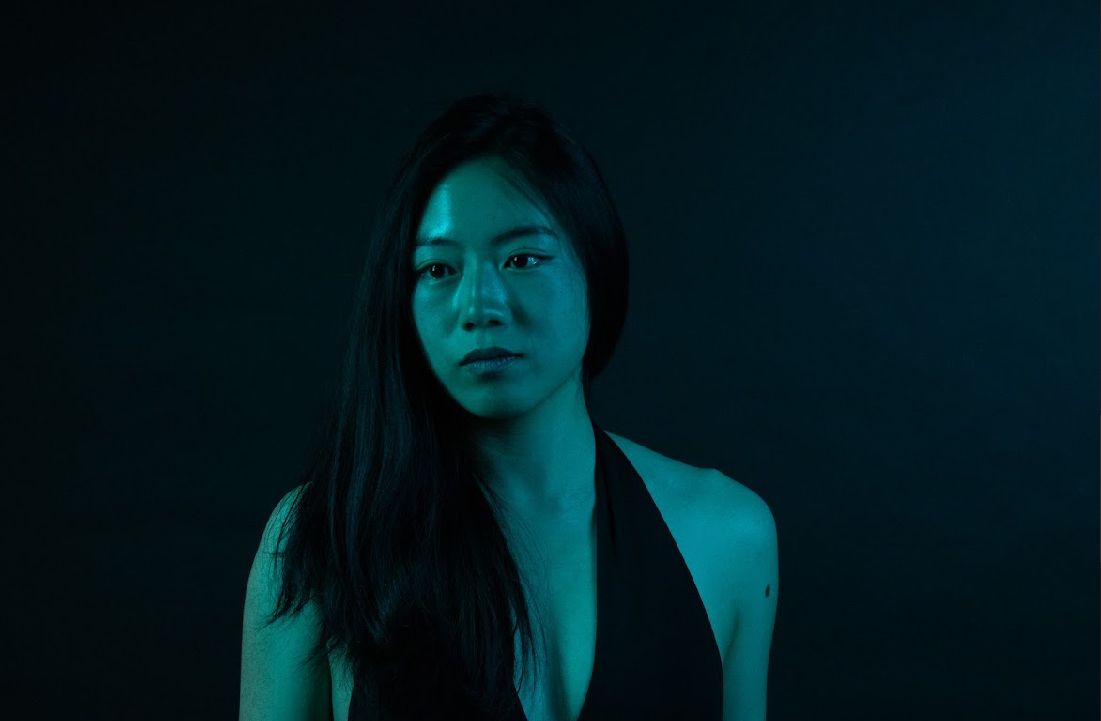 Woman with long dark hair, lit by teal and blue light, looking to the side.