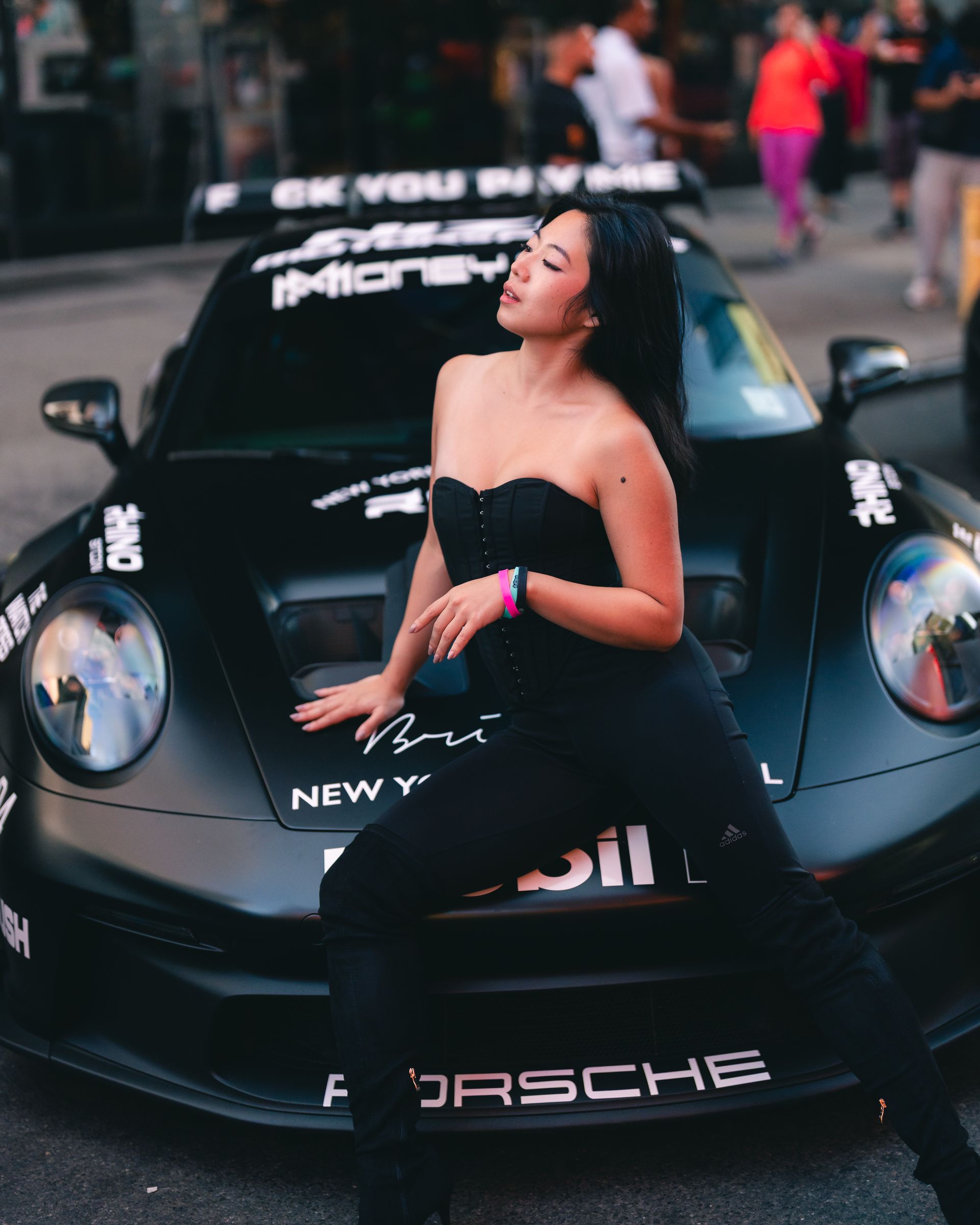 Minnie Lee - NYC model in black outfit poses in front of a black Porsche car,  at One Life Car Rally.