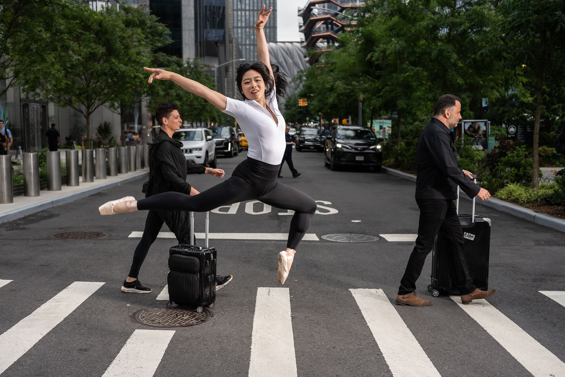 A ballerina leaps in a crosswalk with two people walking alongside, holding suitcases. City background.