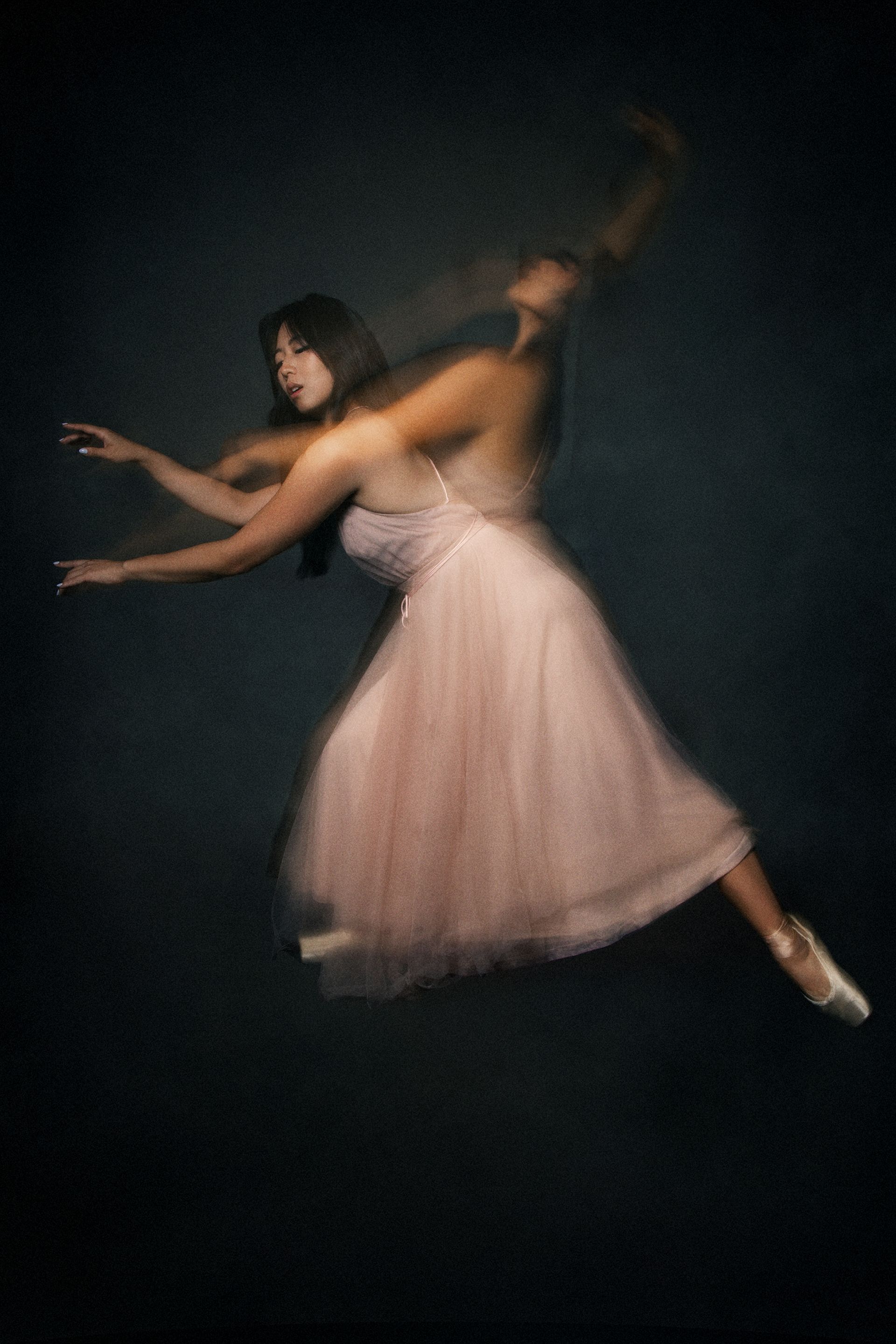 Ballet dancer in pink dress leaps, blurred motion effect against dark background.