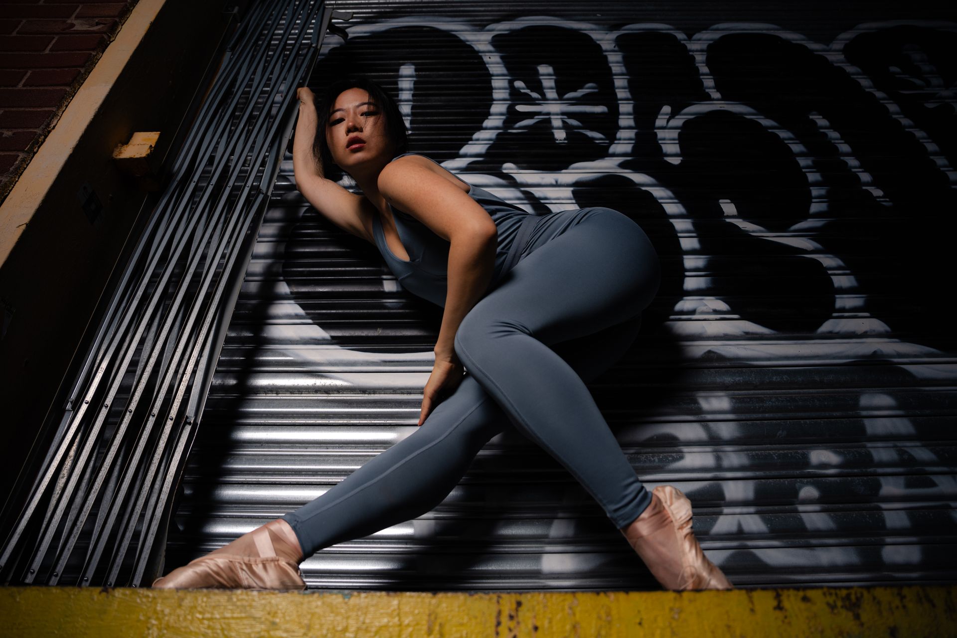 Asian Woman in ballet shoes leans against a graffiti-covered wall in a dramatic pose, wearing a gray jumpsuit.