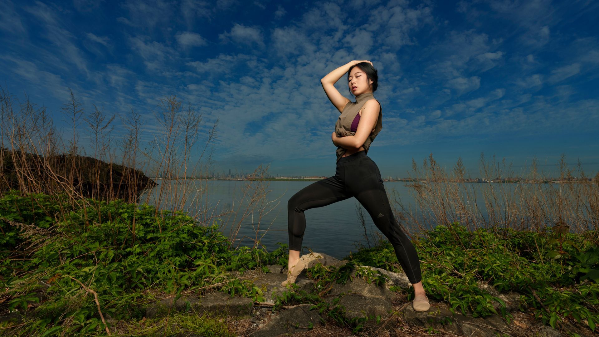 Woman in athletic wear posing outdoors by water, hand on head, blue sky.
