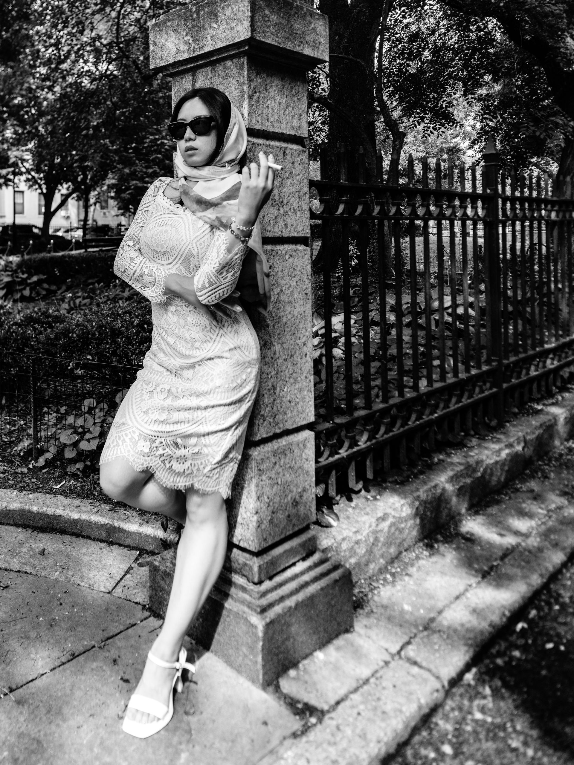 vintage Woman in sunglasses, dress, and scarf, smoking a cigarette, leaning against a stone pillar next to a iron fence.