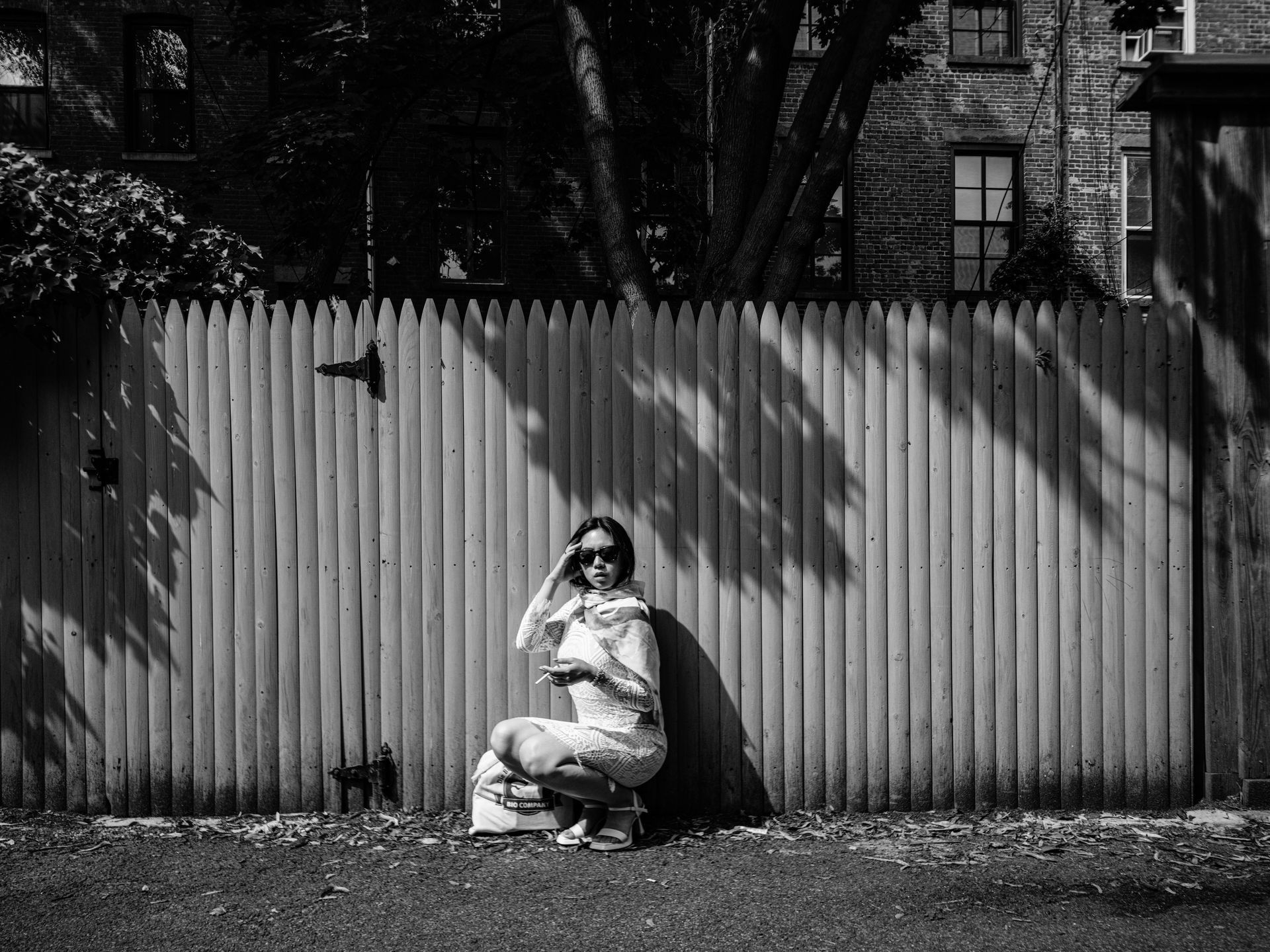 Woman squatting by a wooden fence, wearing sunglasses, holding a cigarette in a sunny backyard.