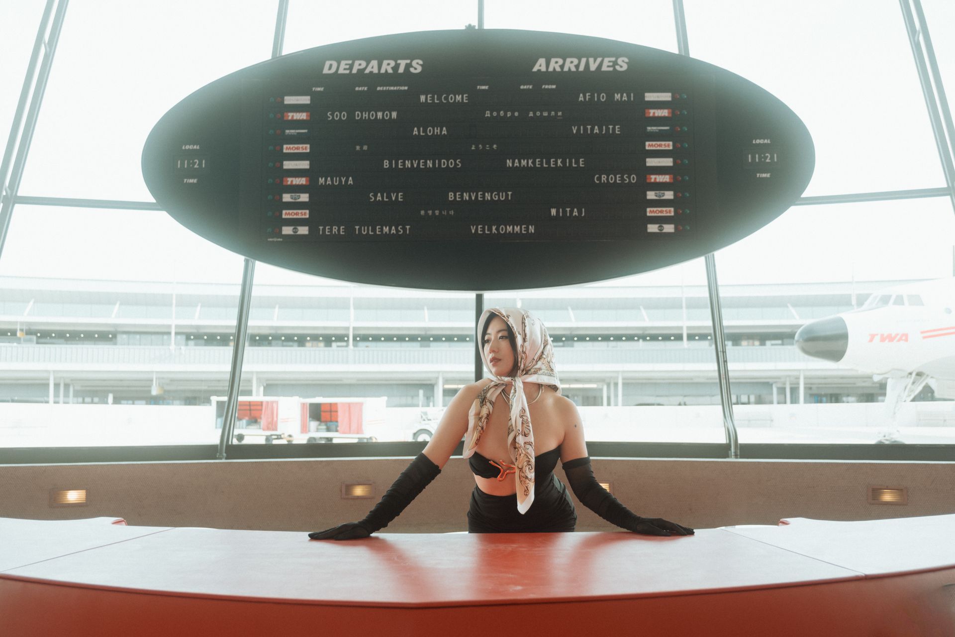 Woman in vintage looking white lace dress and sunglasses and white scarf poses outside near stone wall, hand to forehead.