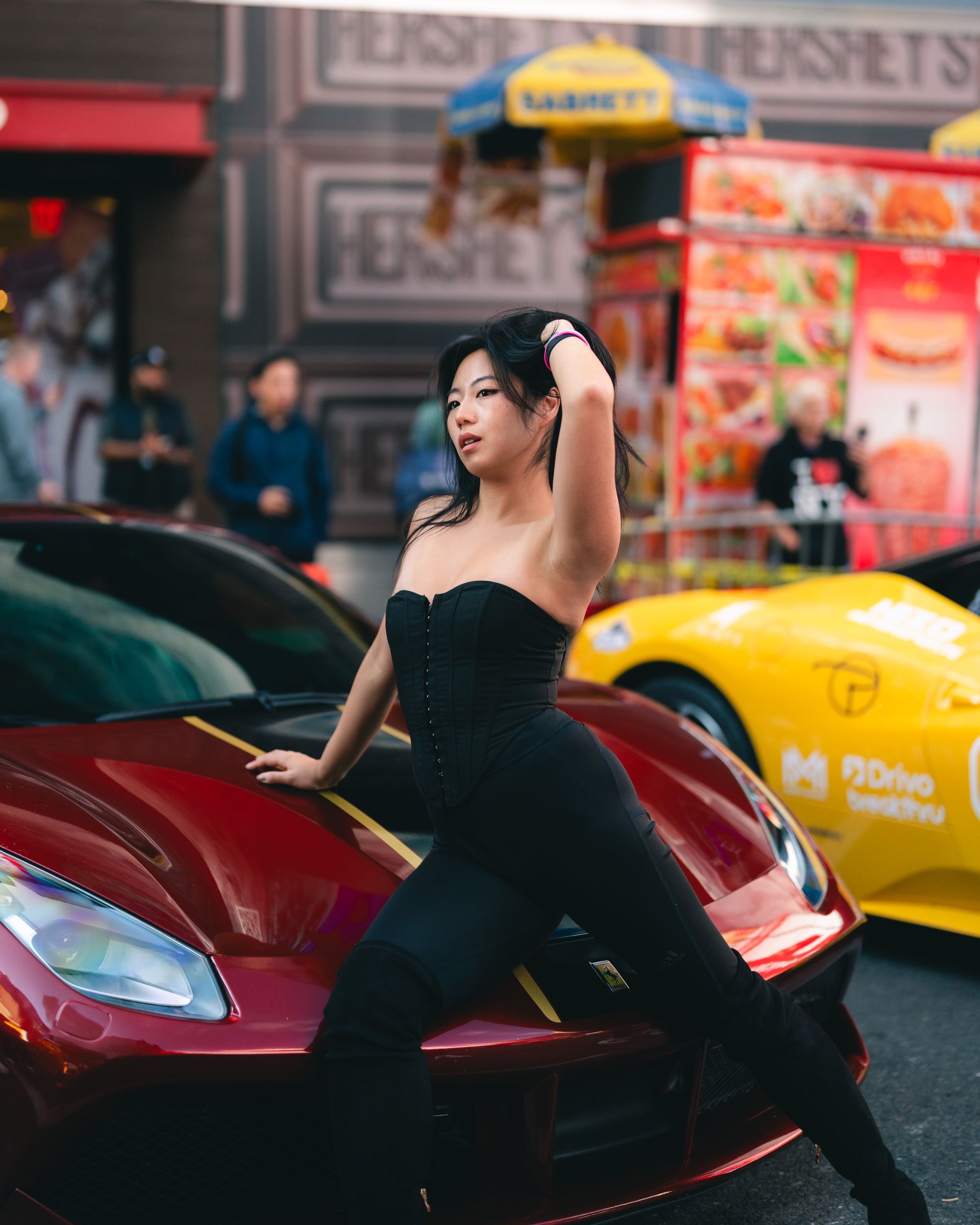 Woman in black outfit poses on red sports car lamborghini, yellow car behind. Street scene with food cart.
