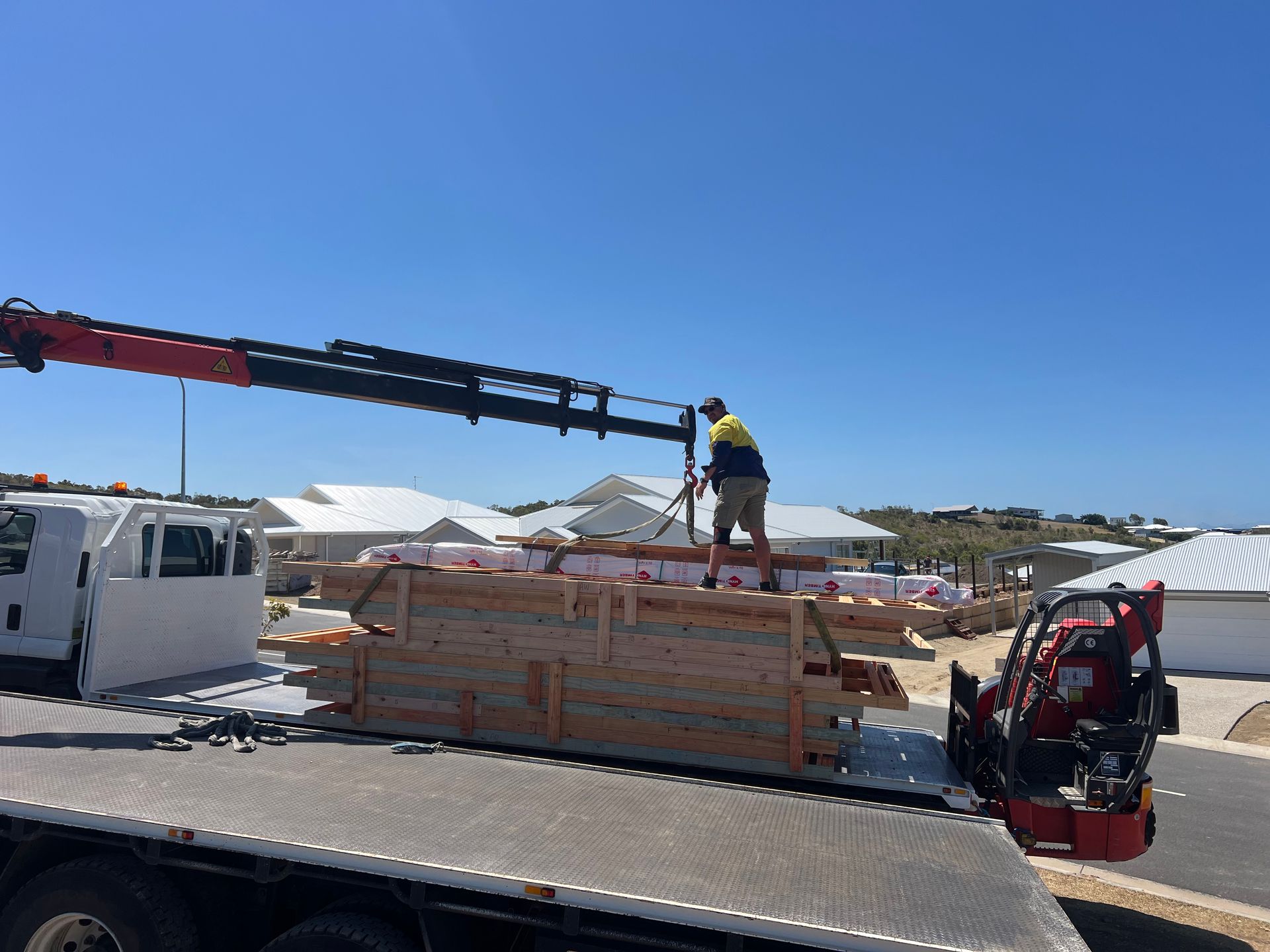 Worker on stacked lumber near a crane truck at a construction site under a clear blue sky