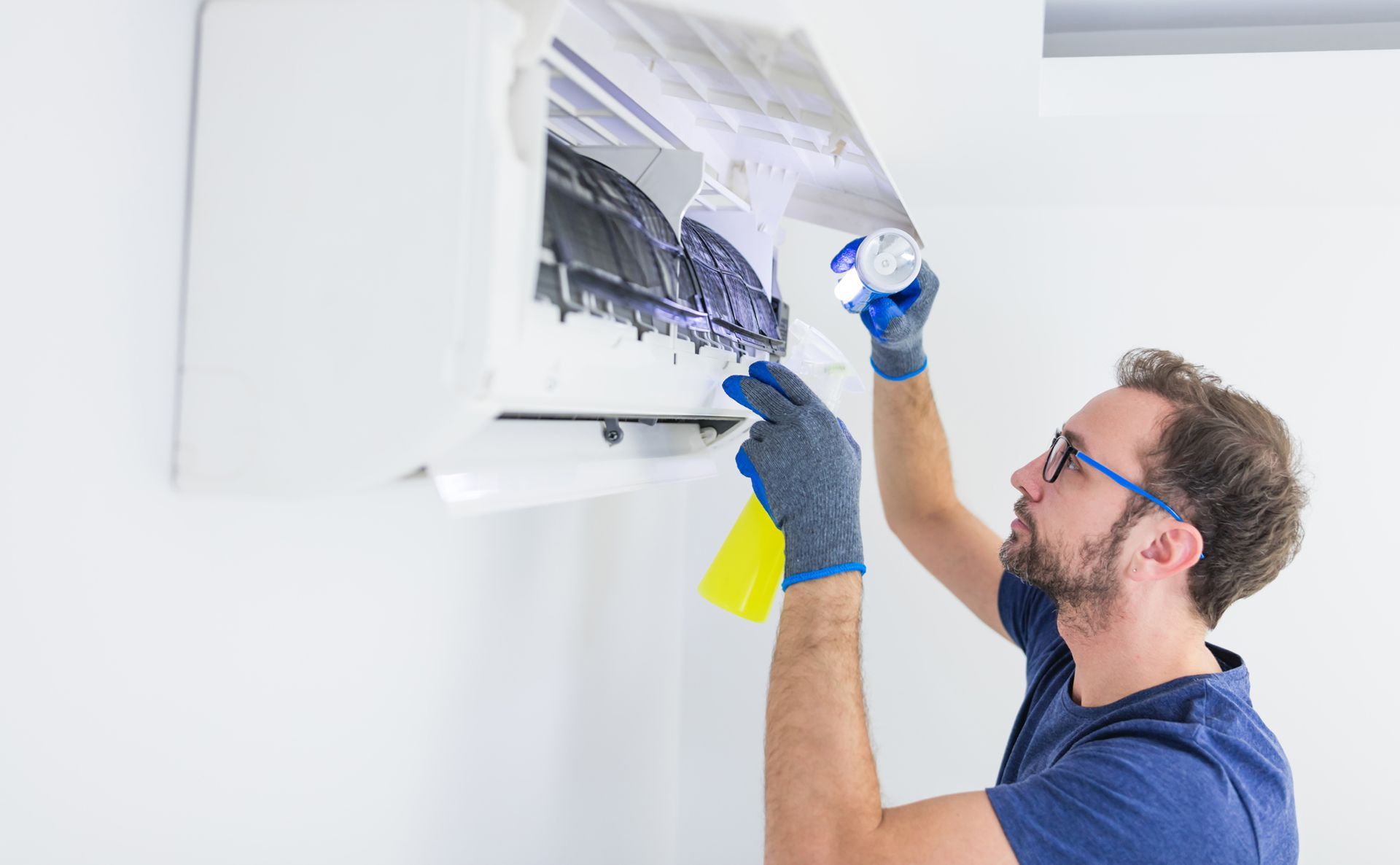 Man cleaning an AC unit with spray bottle and sponge. White wall backdrop.