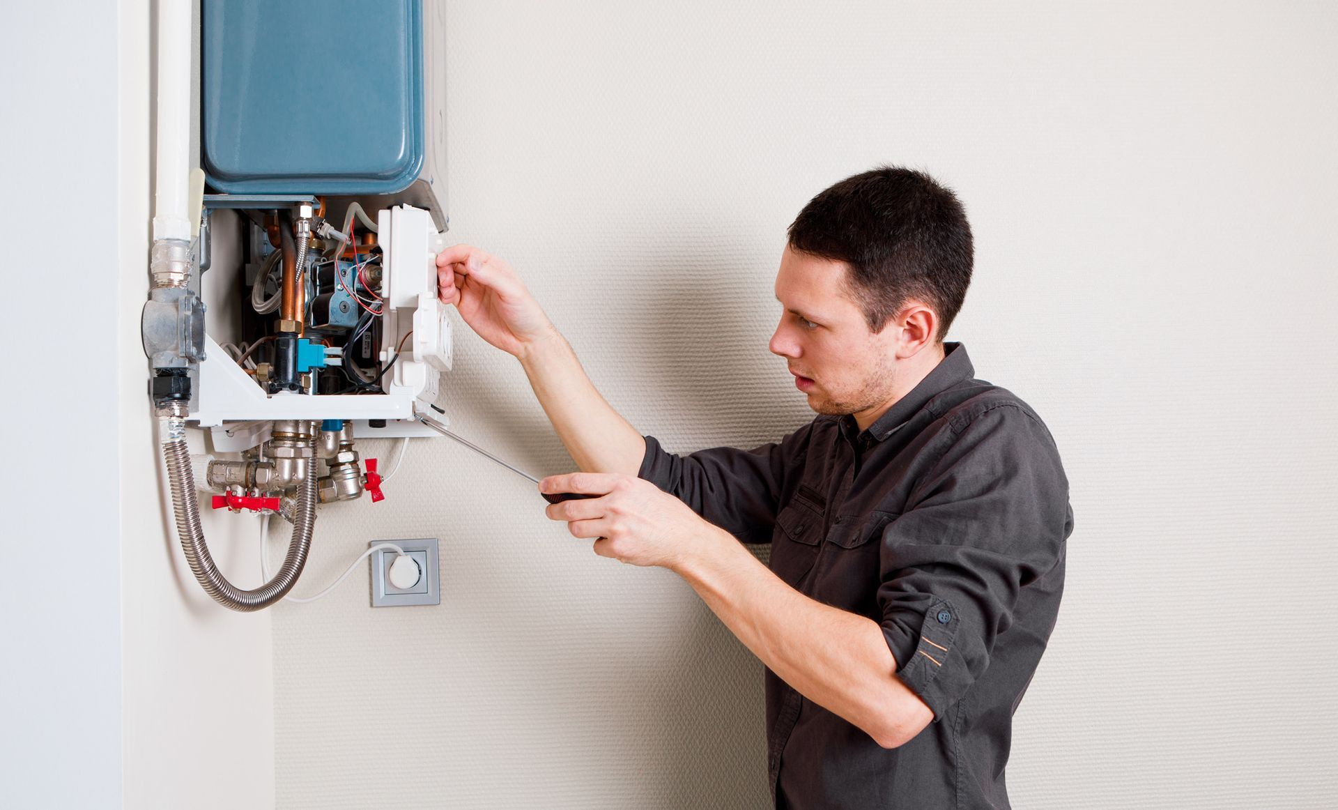 Man using a screwdriver to work on a wall-mounted blue water heater. White wall, indoor setting.