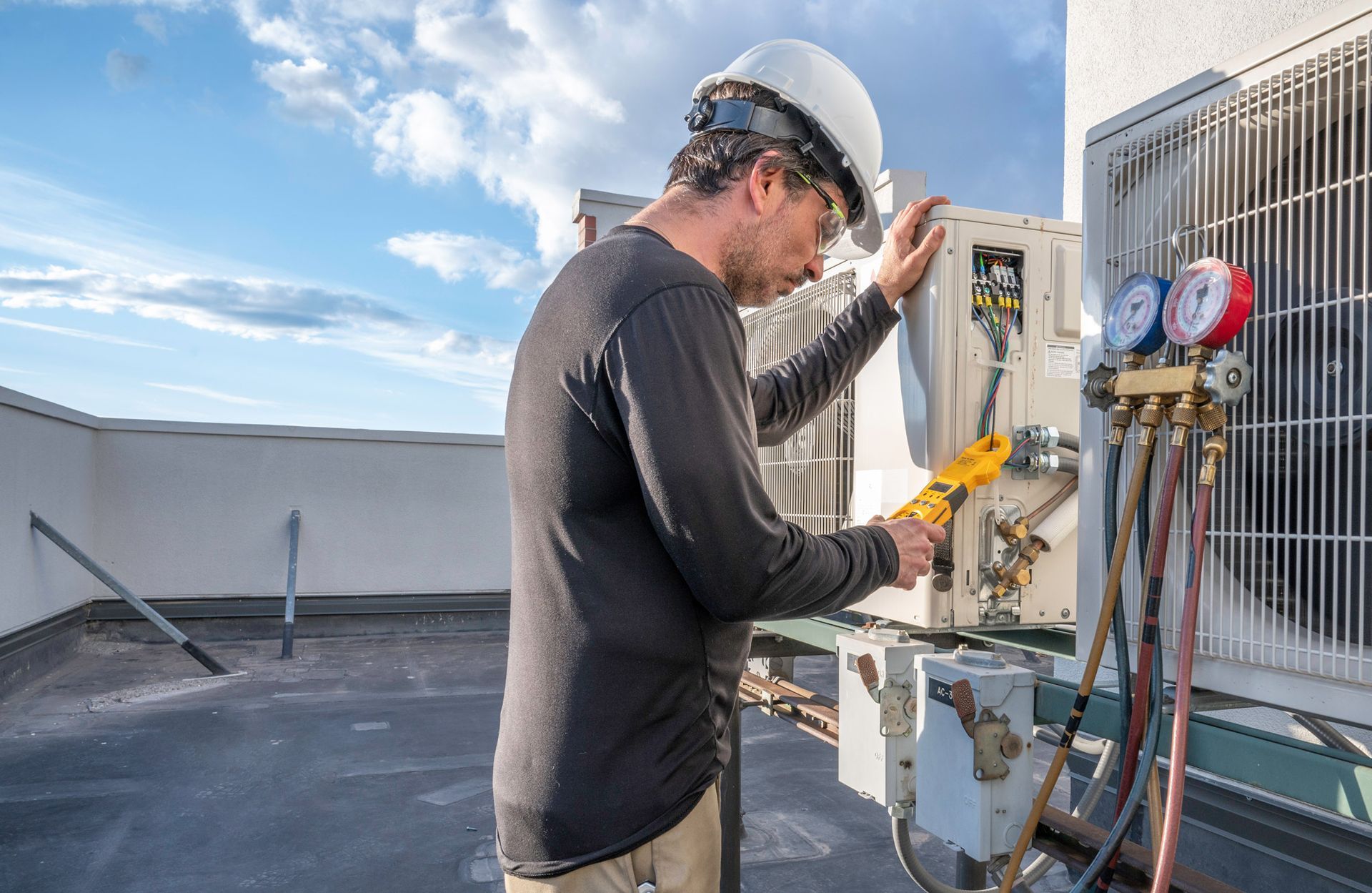 HVAC technician on a rooftop inspects air conditioning unit with gauges. Blue sky.
