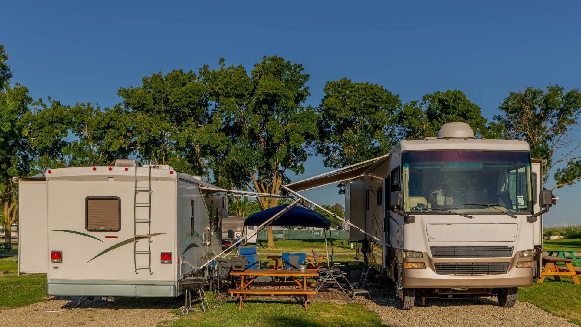 Two RVs parked side-by-side at a campsite. Awnings extended, picnic table, trees, and blue sky.