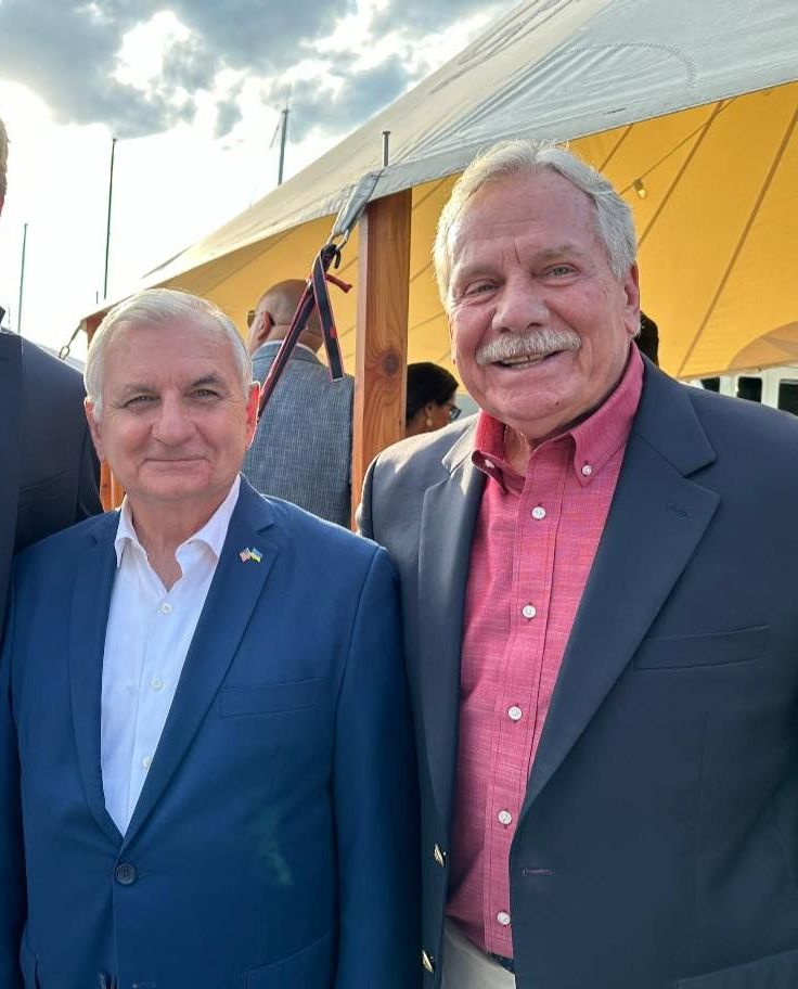 Two men in suits pose for a picture in front of a tent
