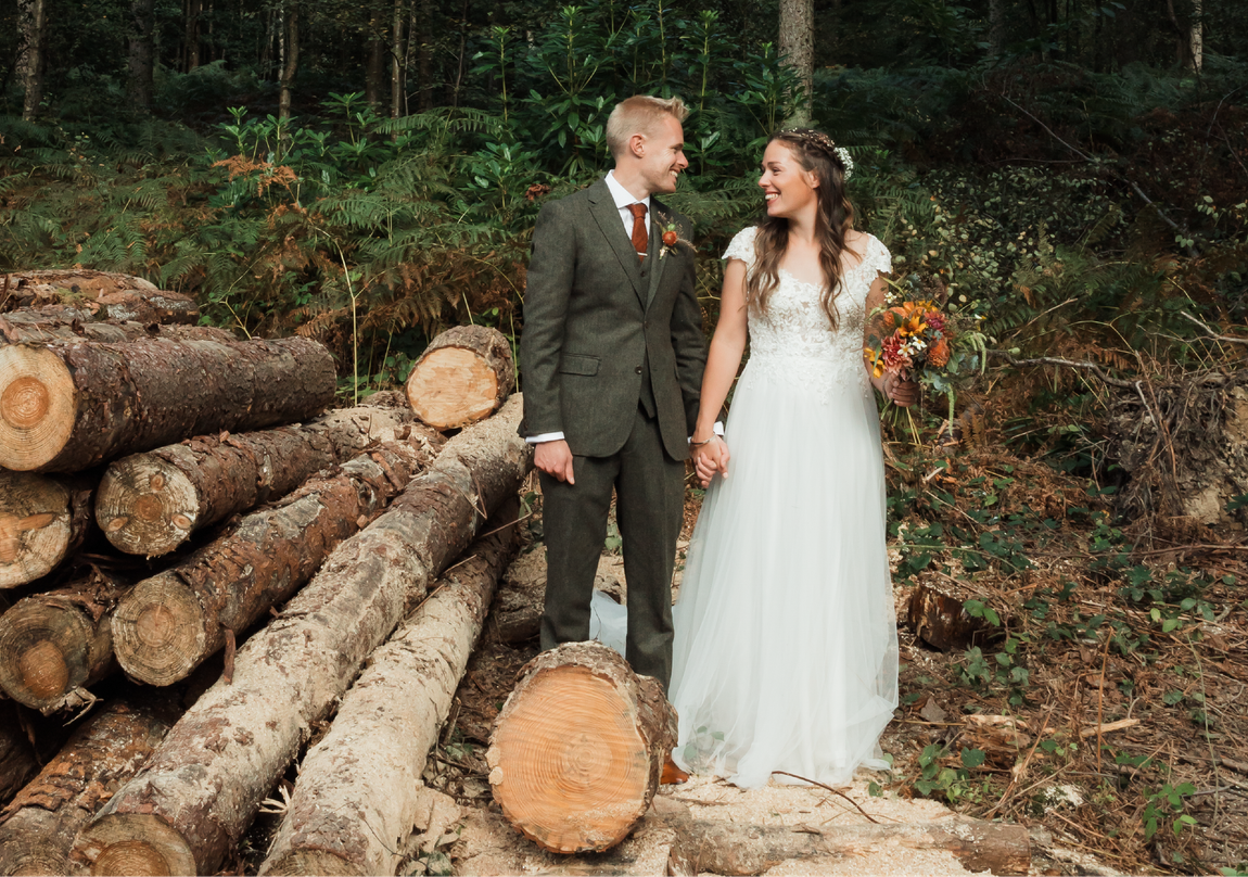 A bride and groom are standing next to a pile of logs in the woods.