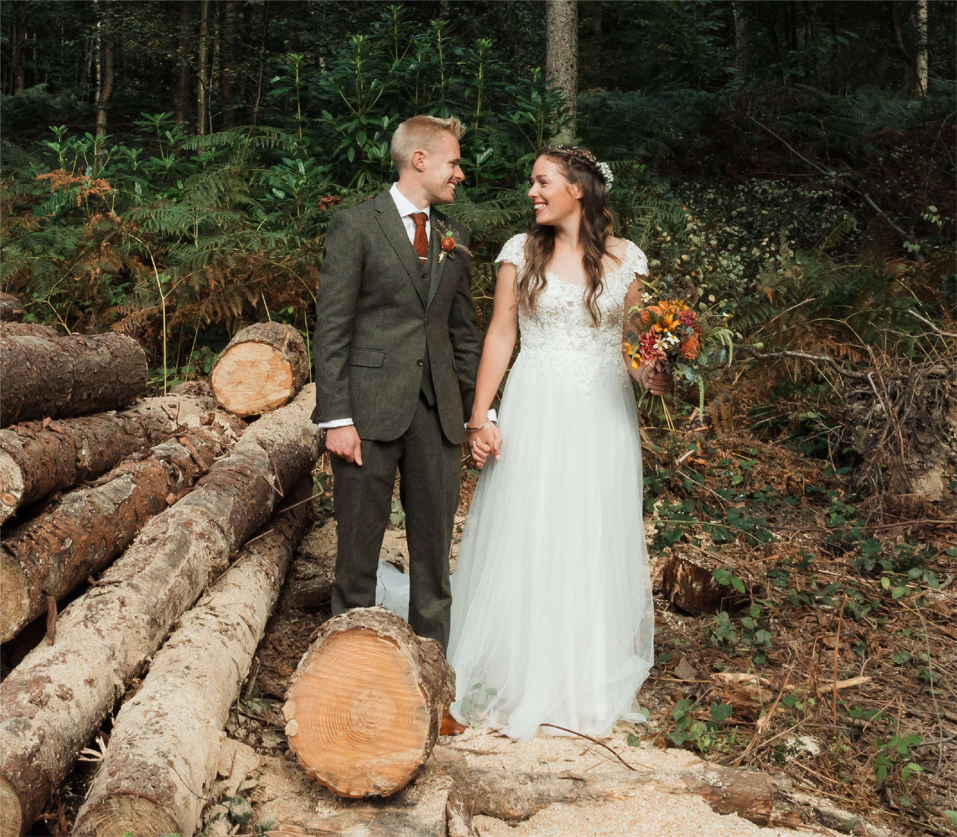 A bride and groom are standing next to a pile of logs in the woods.