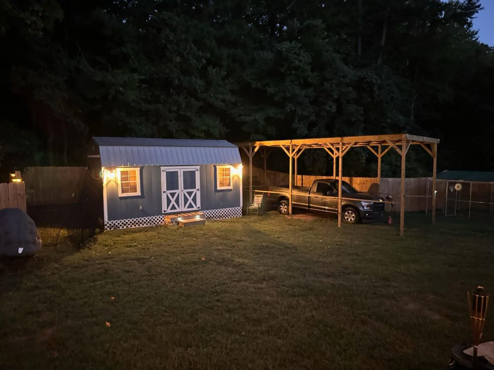 A truck is parked under a carport next to a shed at night.