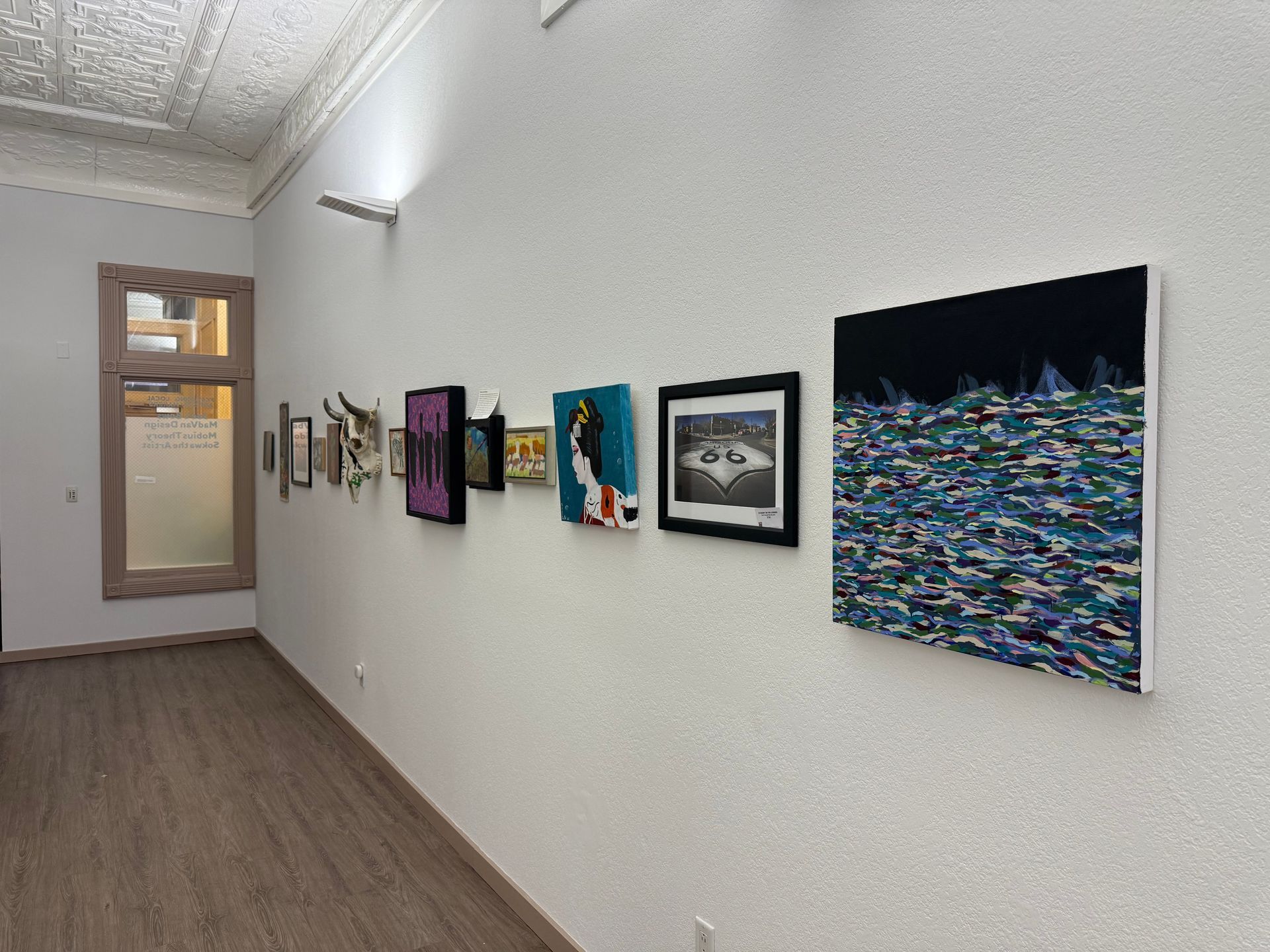 A gallery hallway with a series of diverse framed and canvas artworks hanging along a white wall.