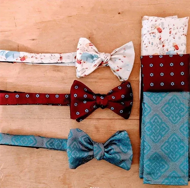 Three bow ties with matching pocket squares in white with red patterns, dark red with blue flowers, and light blue damask.