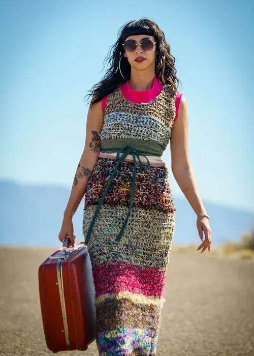 A person in a multi-colored crochet dress and sunglasses walks down a desert road, carrying a vintage brown suitcase.
