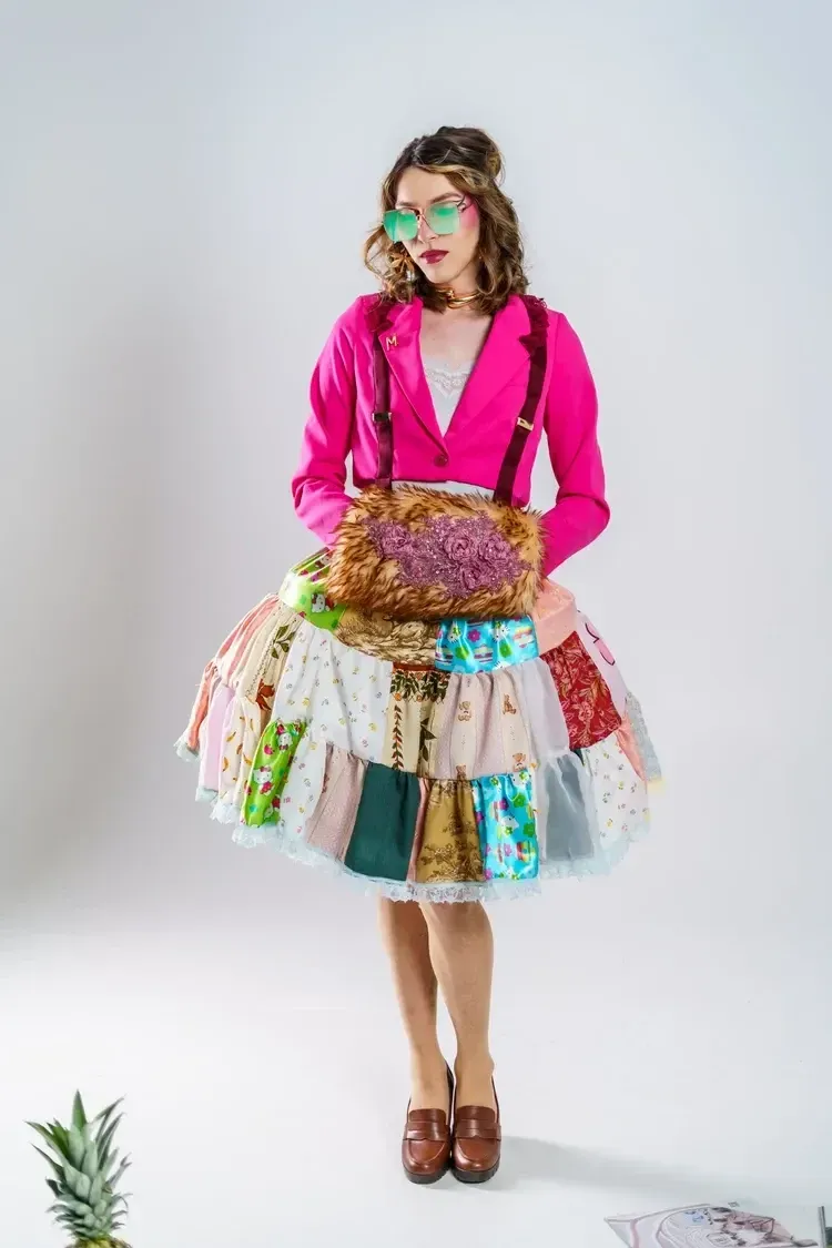 A person wearing bright green glasses, a hot pink jacket, and a colorful, layered patchwork skirt stands against a wall.