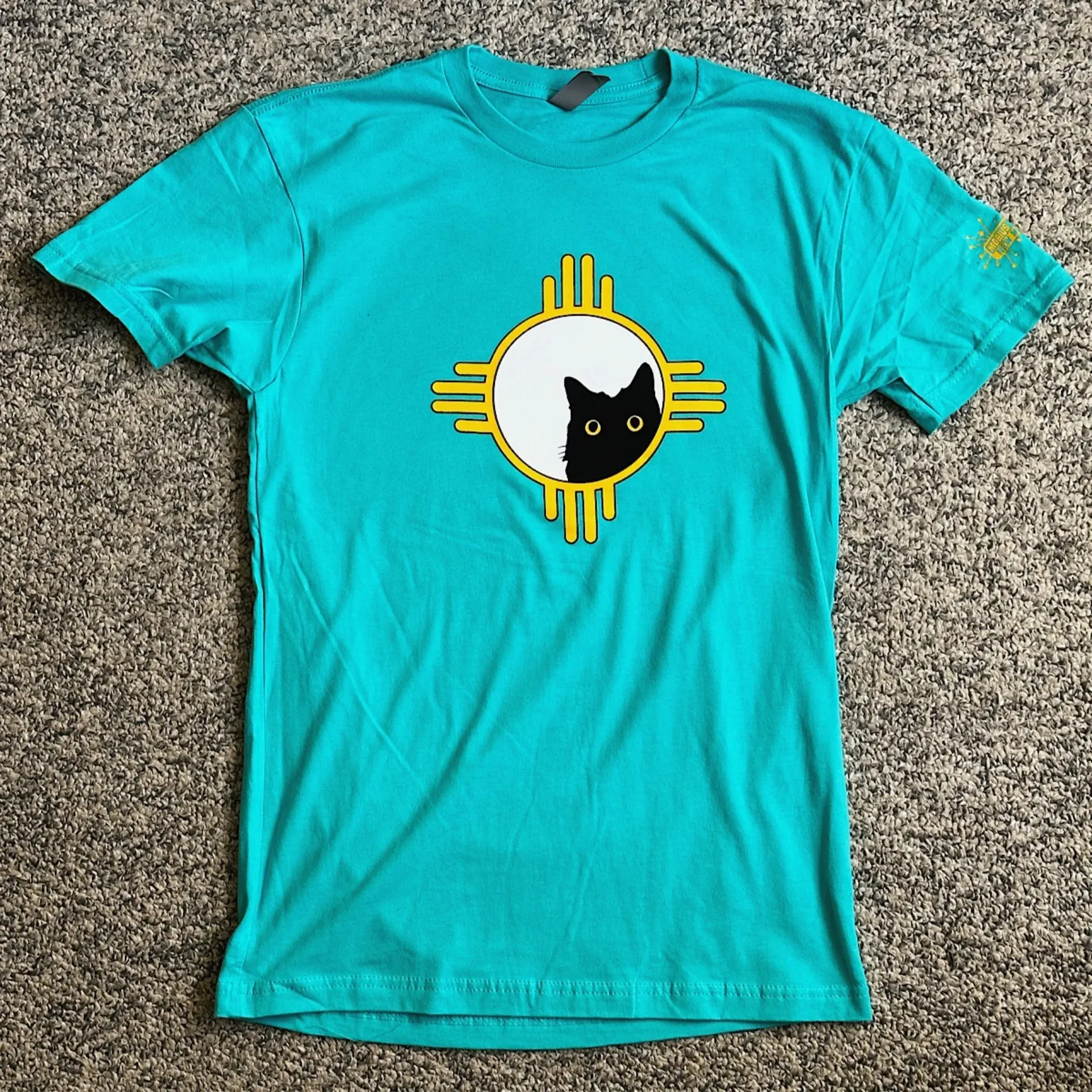 A teal t-shirt featuring a yellow New Mexico sun symbol with a black cat peering from the center circle.