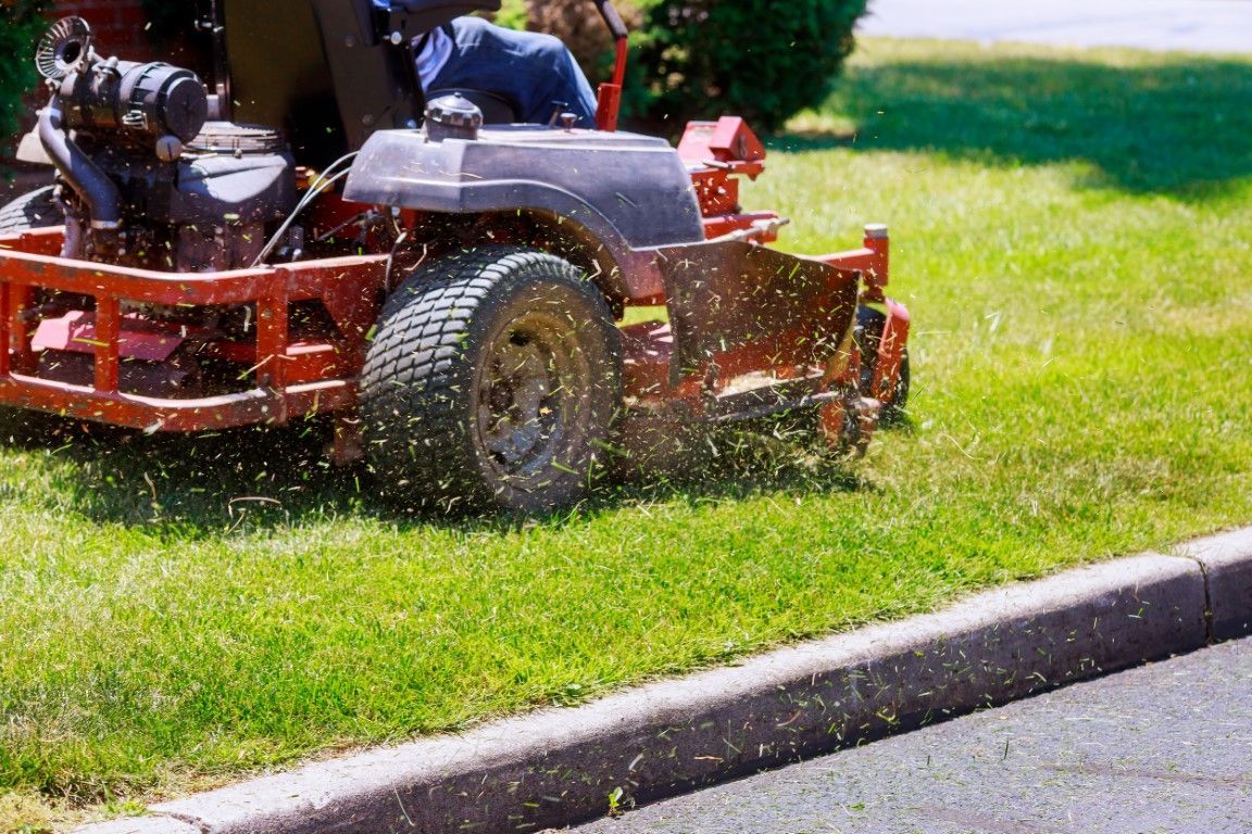 Person operating a red riding lawn mower cutting grass near a curb, with grass clippings flying in the sunlight.