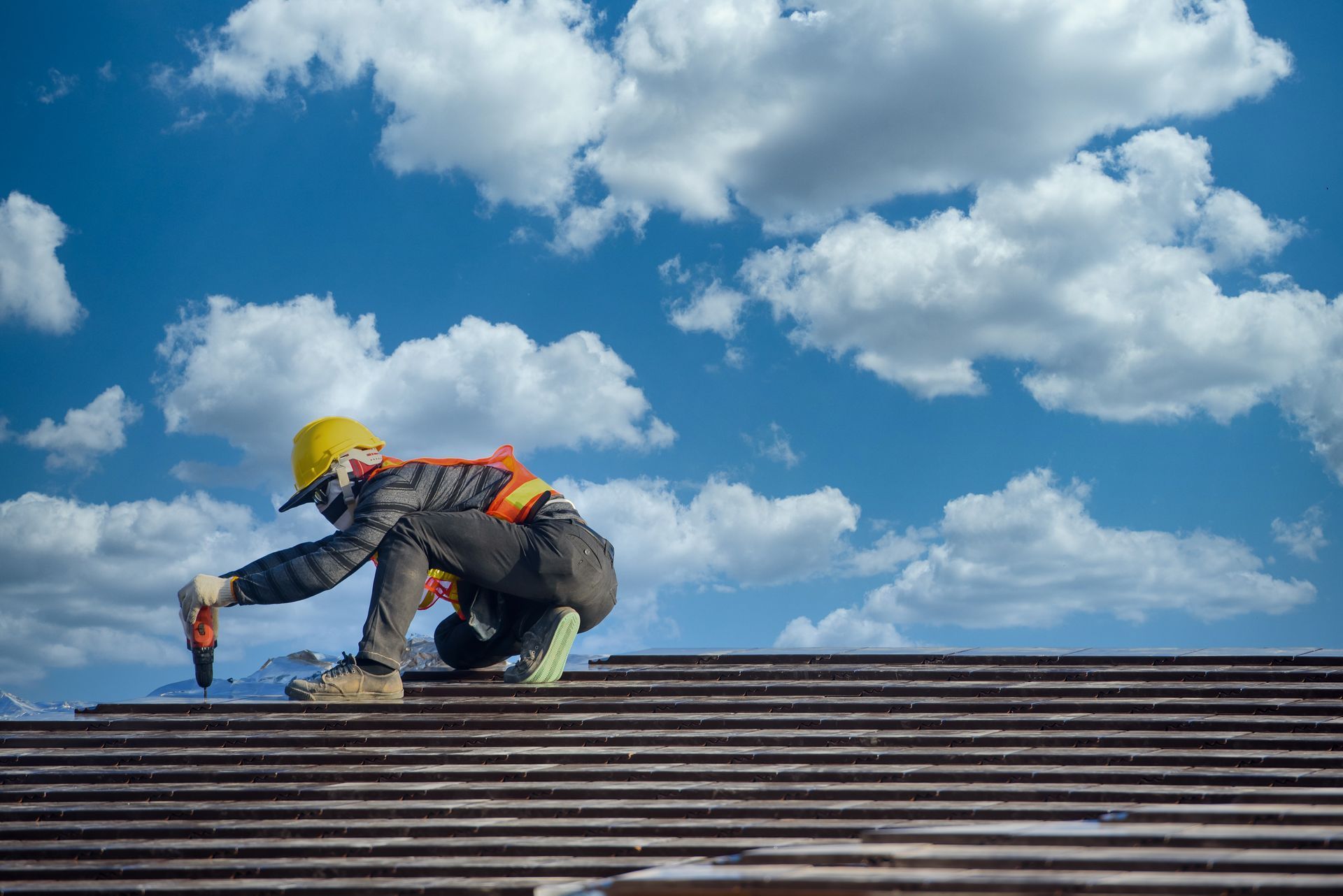 Roofer in safety gear installing shingles on a roof under a blue sky with fluffy clouds.