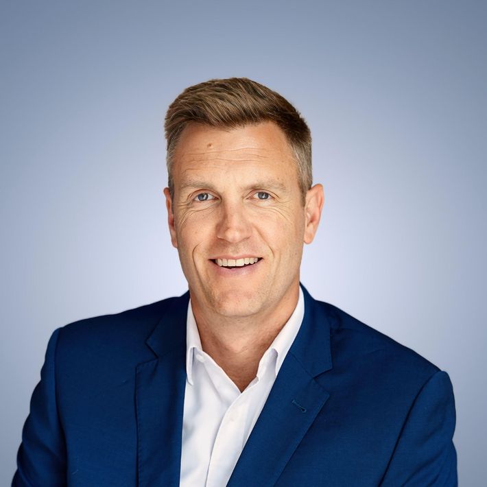 Smiling man in a blue blazer and white shirt, set against a blue gradient background.