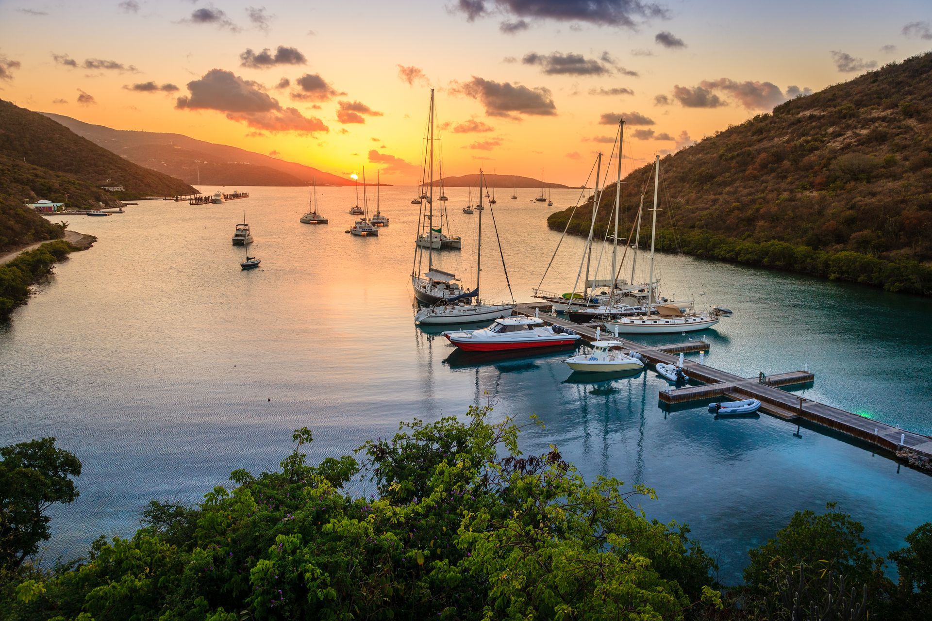 Sunset over a harbor with sailboats moored at docks. Golden light reflects on the water, hills, and sky.