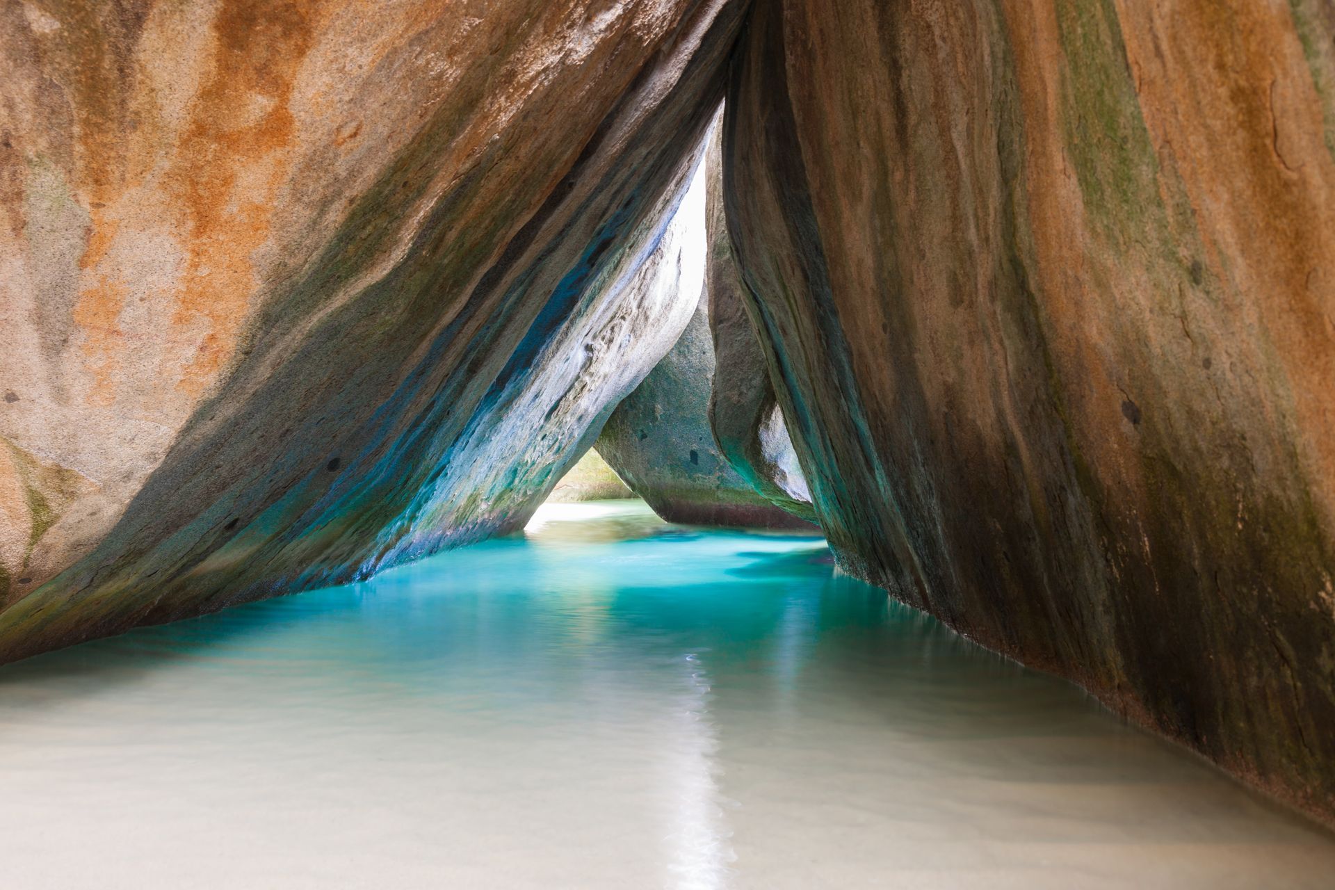 Turquoise water flows through a narrow passage between large, rounded granite boulders; sunlit opening visible.