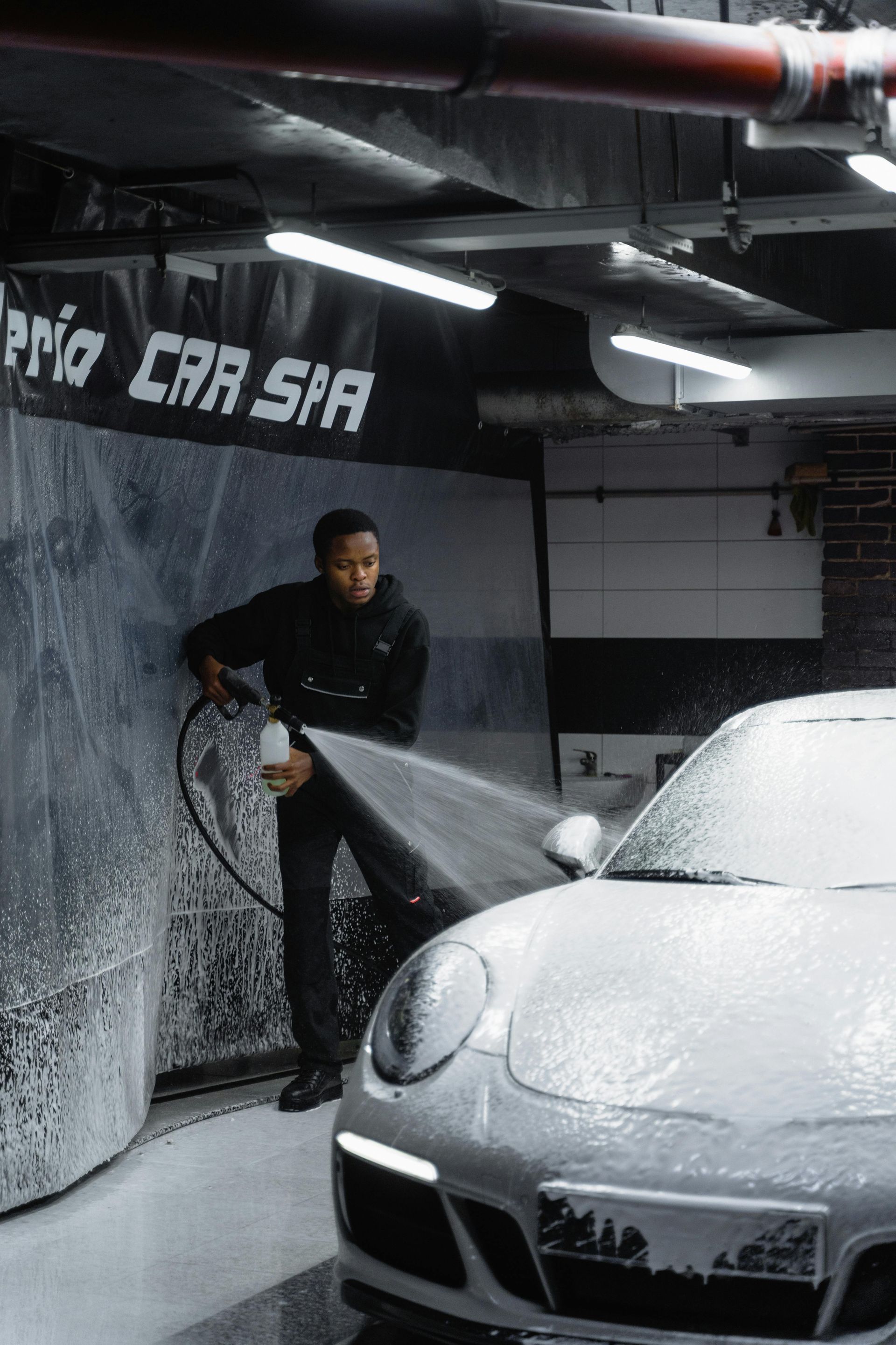 Person washing a silver car with a foam cannon inside a car wash bay.