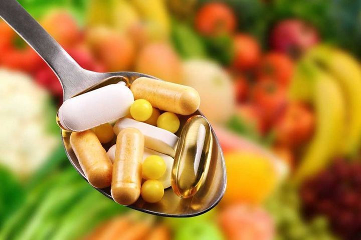 A spoon filled with pills and capsules in front of fruits and vegetables.