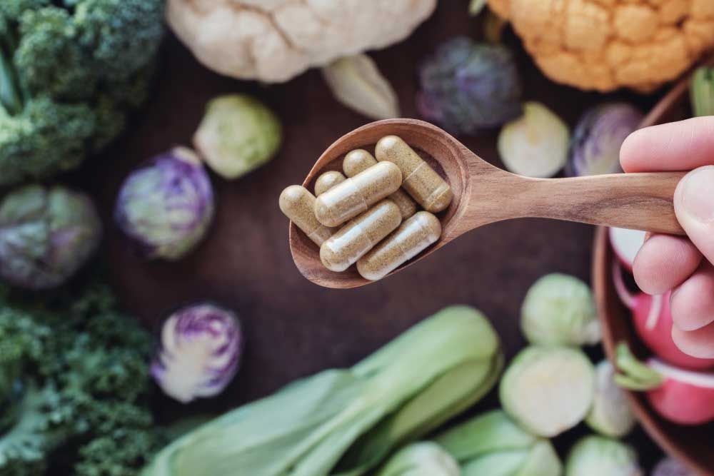 A person is holding a wooden spoon filled with capsules in front of vegetables.