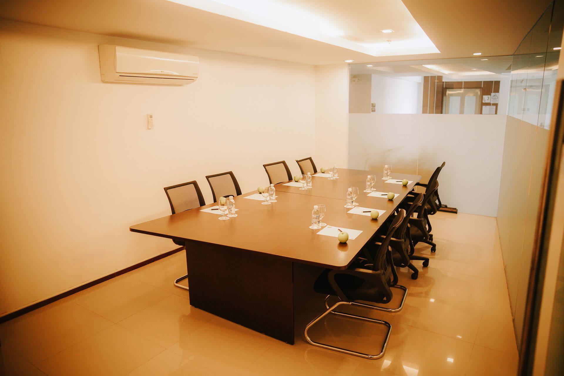 A modern, empty meeting room featuring a rectangular dark wood table surrounded by black office chairs on a light floor.