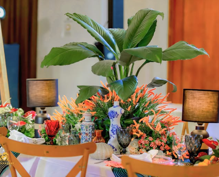 Table setting with large green plant centerpiece, orange flowers, lamps, and blue and white vases.