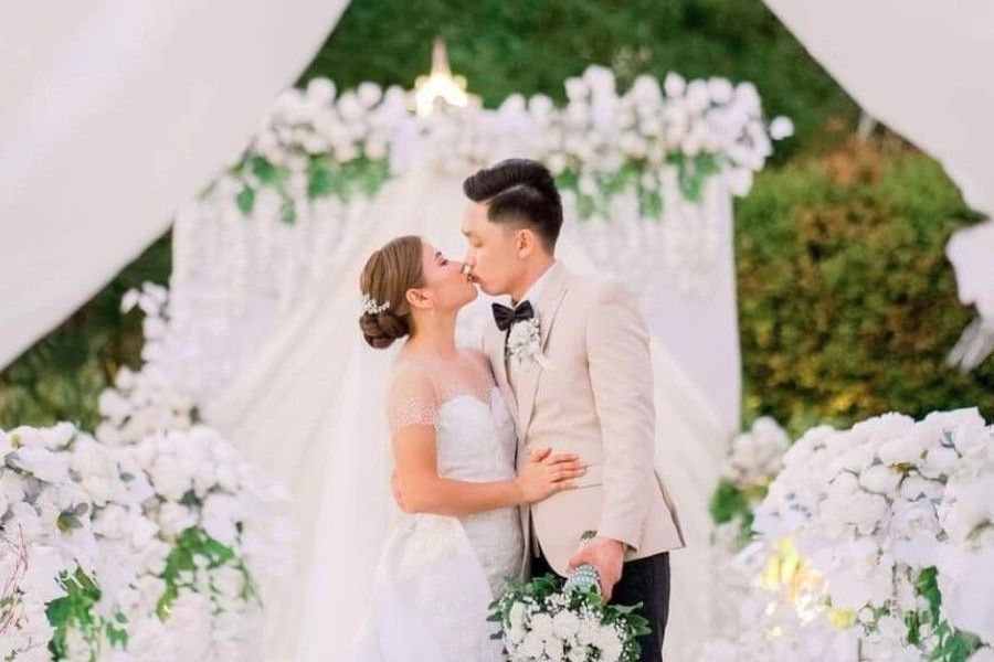 A bride and groom are kissing in front of a white arch.