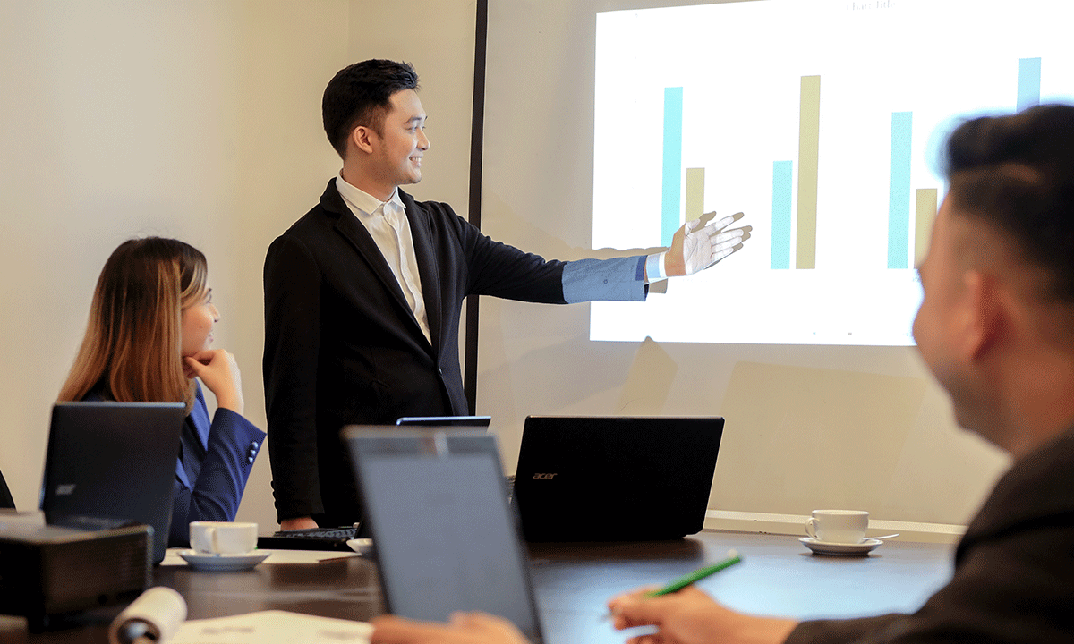 A professional presenting data on a screen to colleagues in a conference room with laptops on the table.
