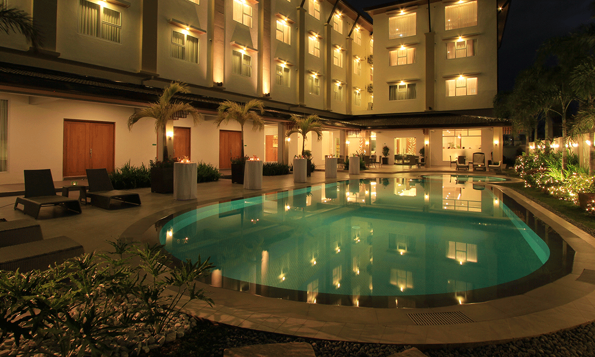 Nighttime view of a hotel pool, reflecting lights from the building. Lounge chairs sit nearby.