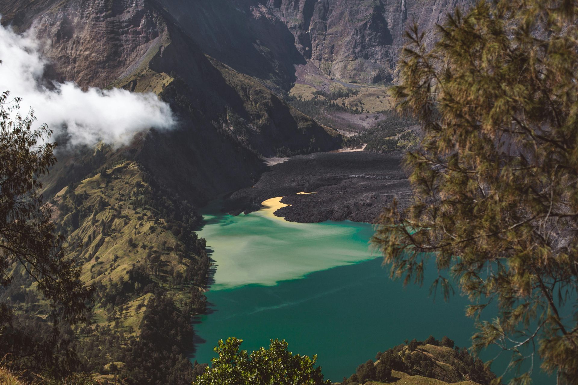 Emerald lake surrounded by dark mountains with clouds, view through trees.