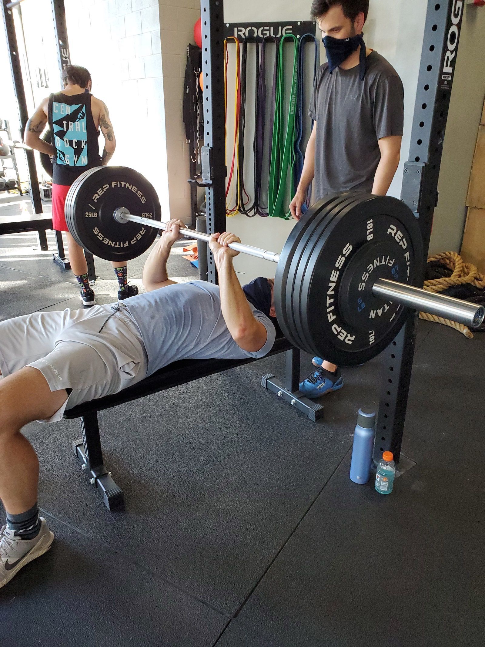 Man bench pressing with spotter in a gym. He's wearing a mask and gym clothes.