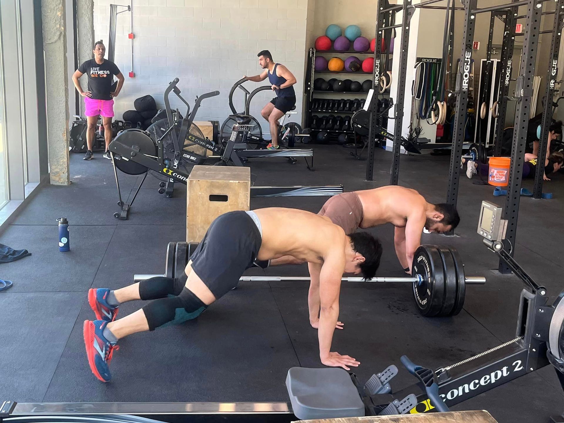 People exercising in a gym: men doing pushups, one on bike, others with weights. Black and gray equipment.
