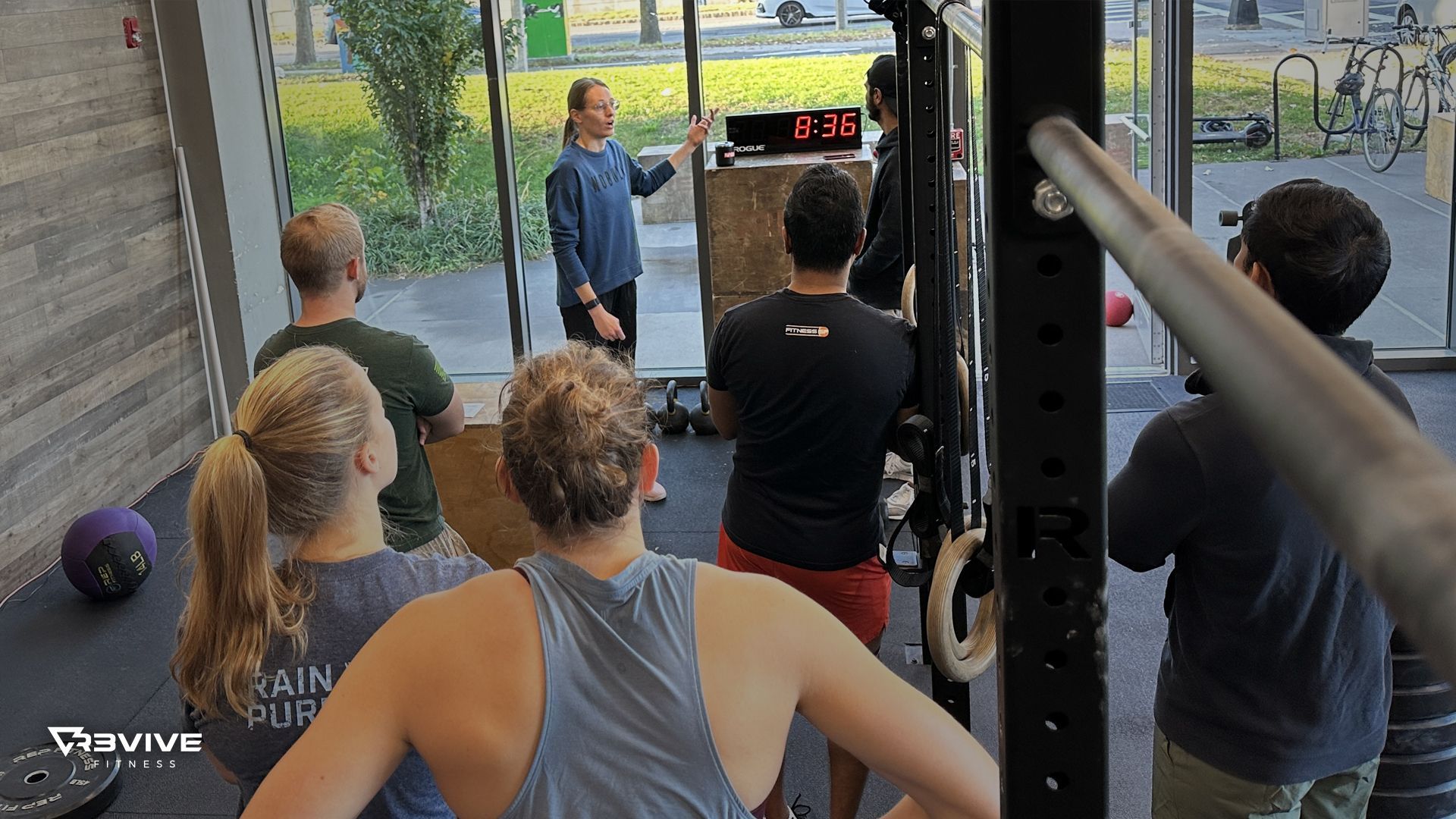 People in gym watching trainer with timer on a box, some wearing athletic clothes, light streaming in from the window.