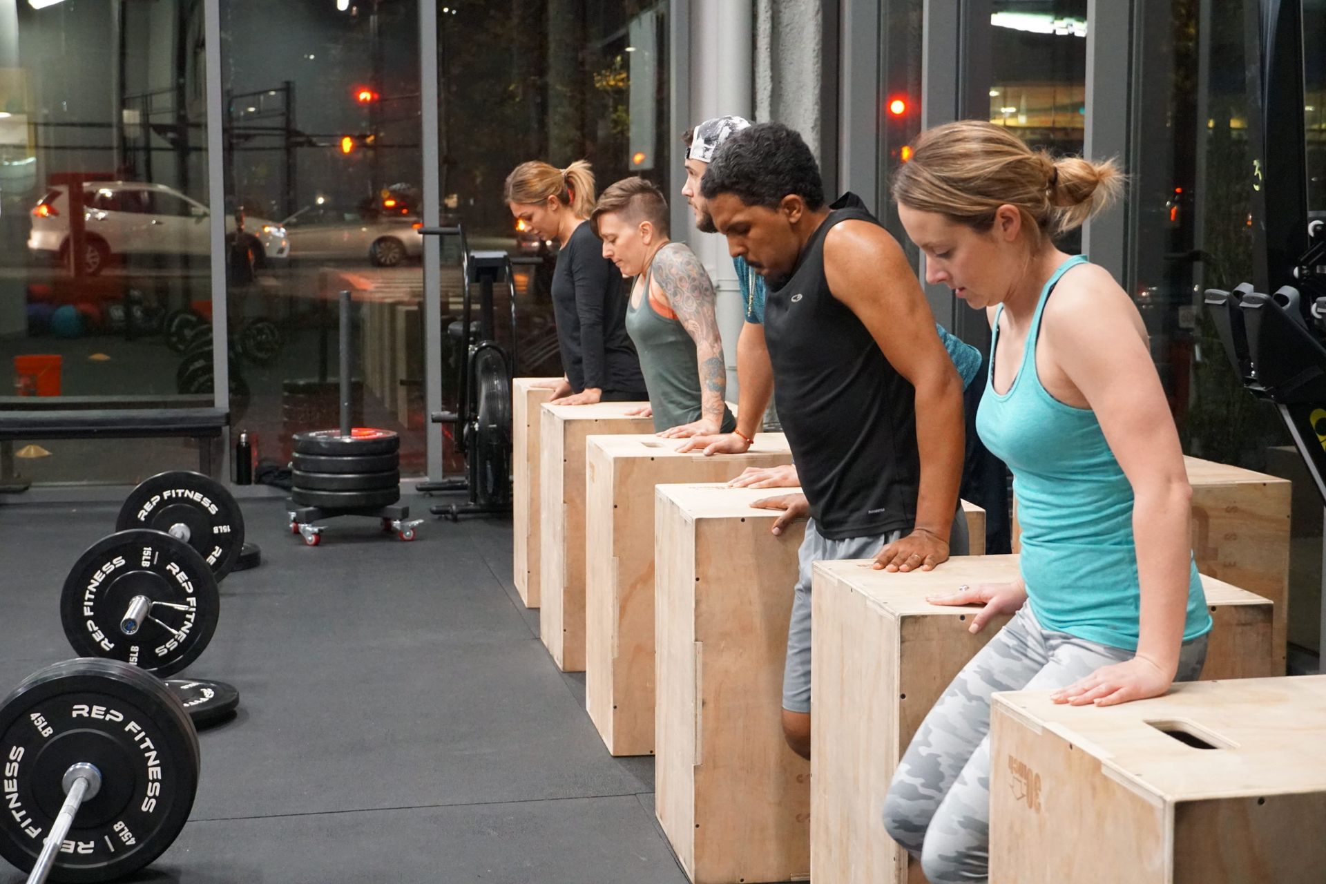 People doing dips on boxes in a gym, with weights in the foreground and city lights visible through windows.