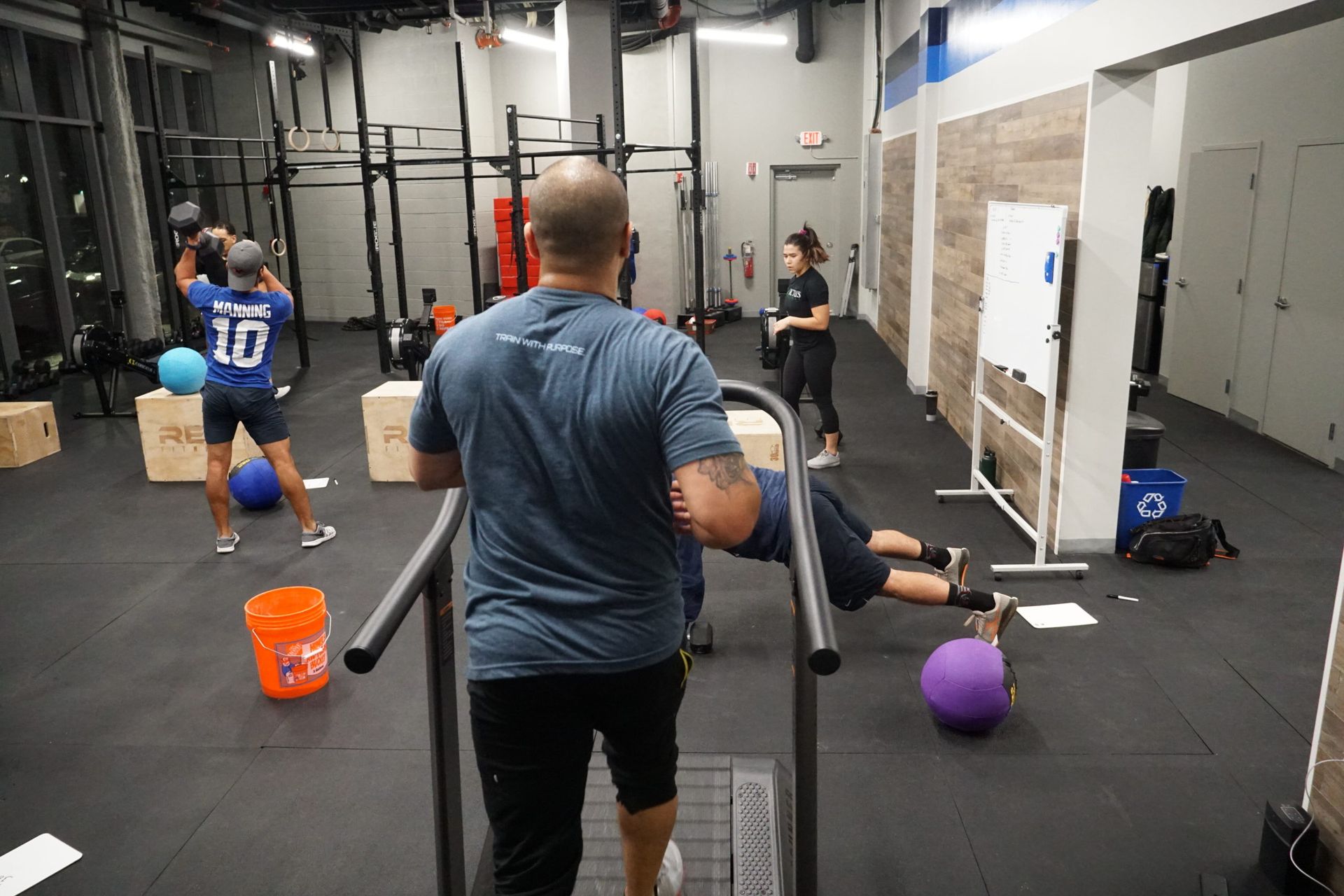 People working out in a gym. A man coaches another as he performs a push-up. Others use weights and equipment.
