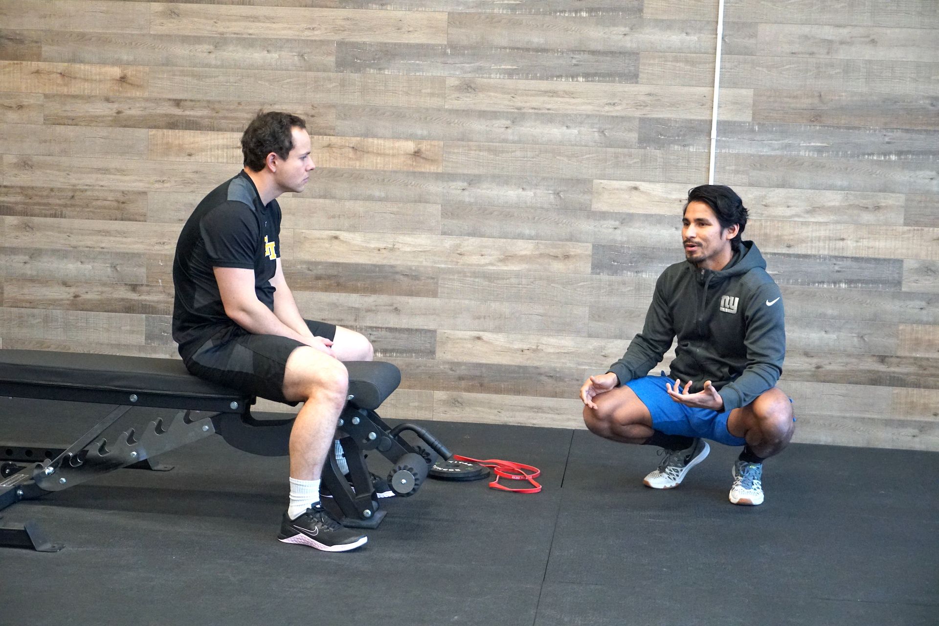 Two men in a gym setting, one sitting on a bench, other squatting, talking.
